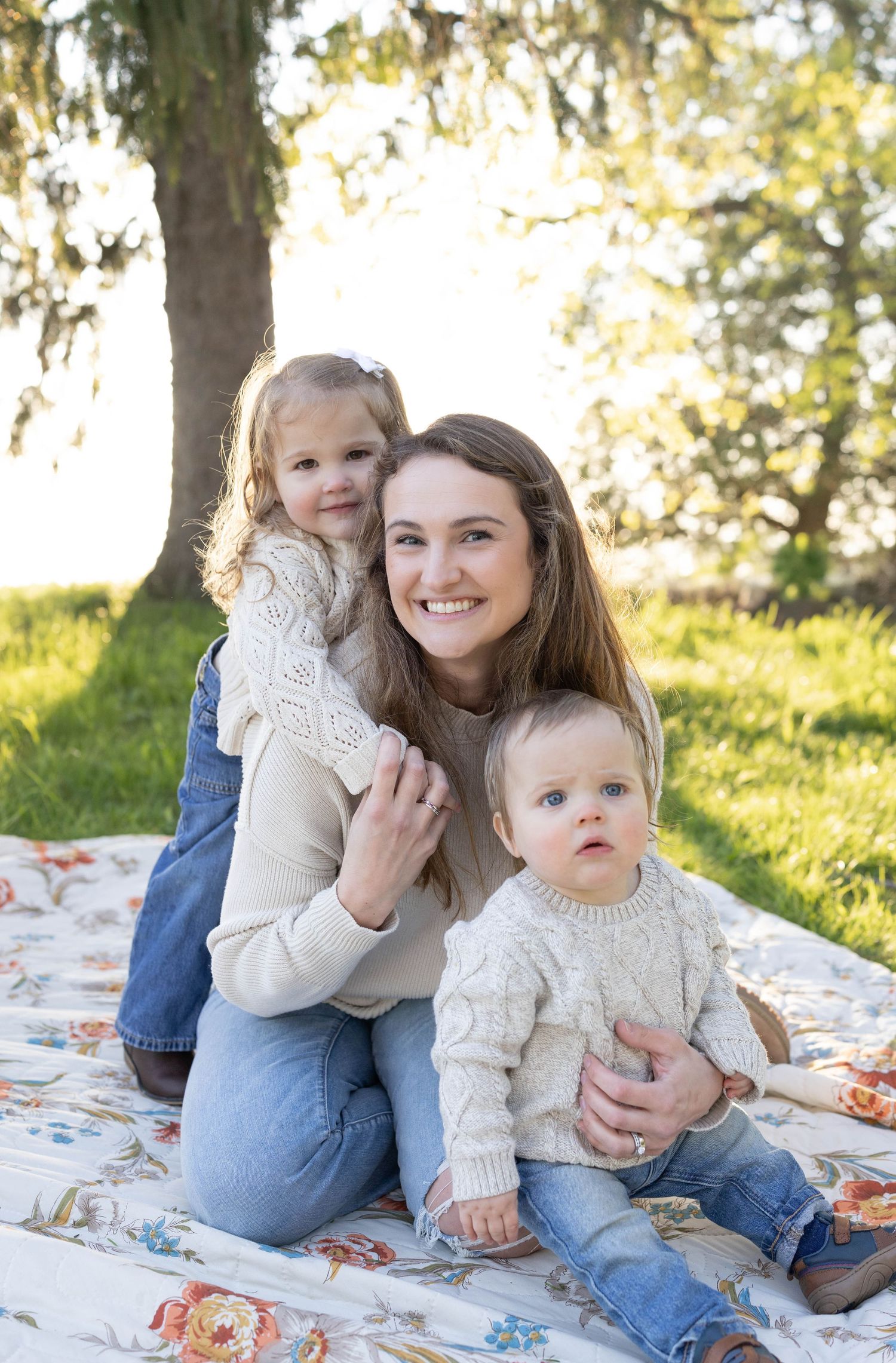 Family enjoying a sunny afternoon together while sitting on a floral blanket in a grassy park setting.