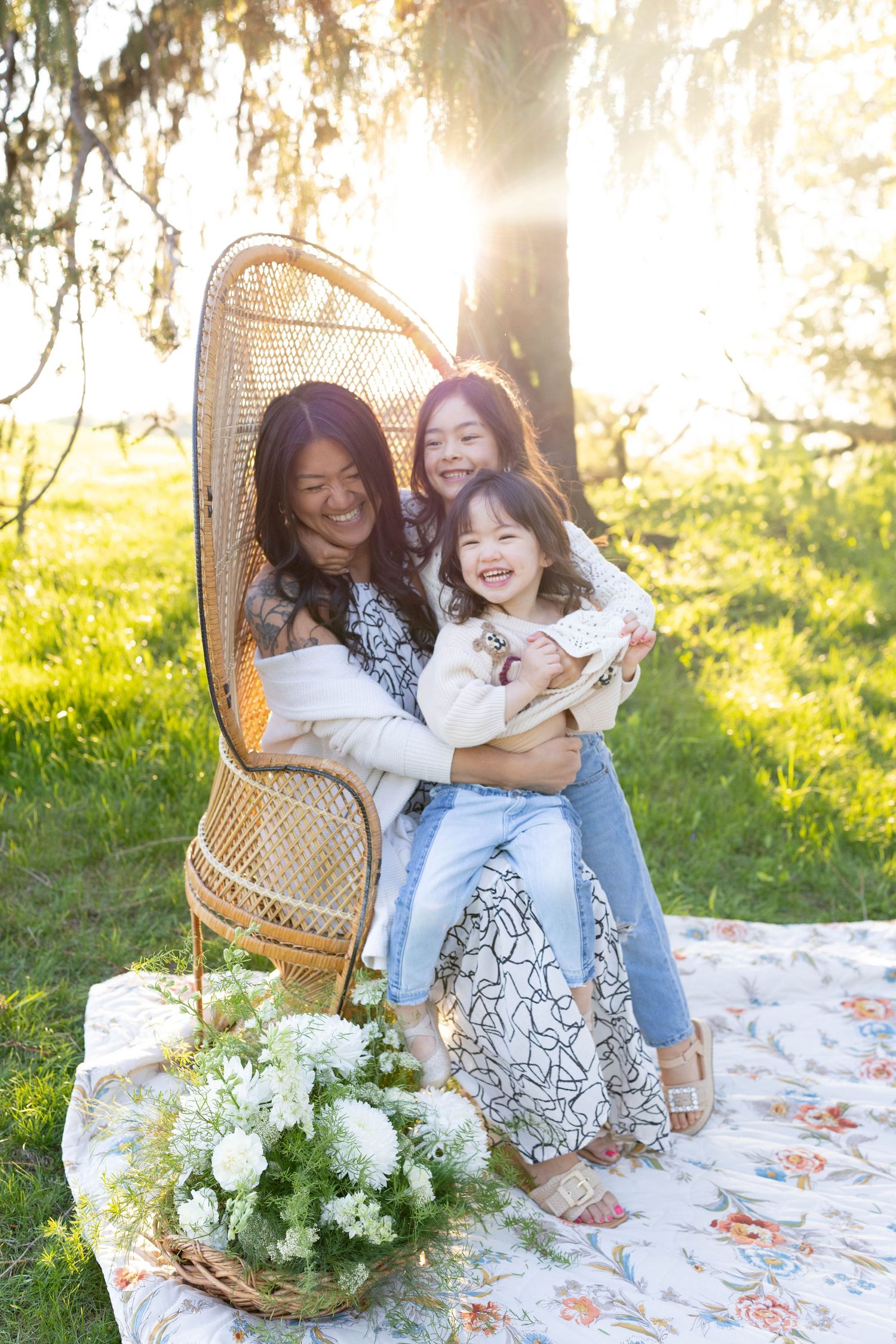 Three people share joyful laughter while sitting together in a wicker peacock chair on a picnic blanket at sunset.