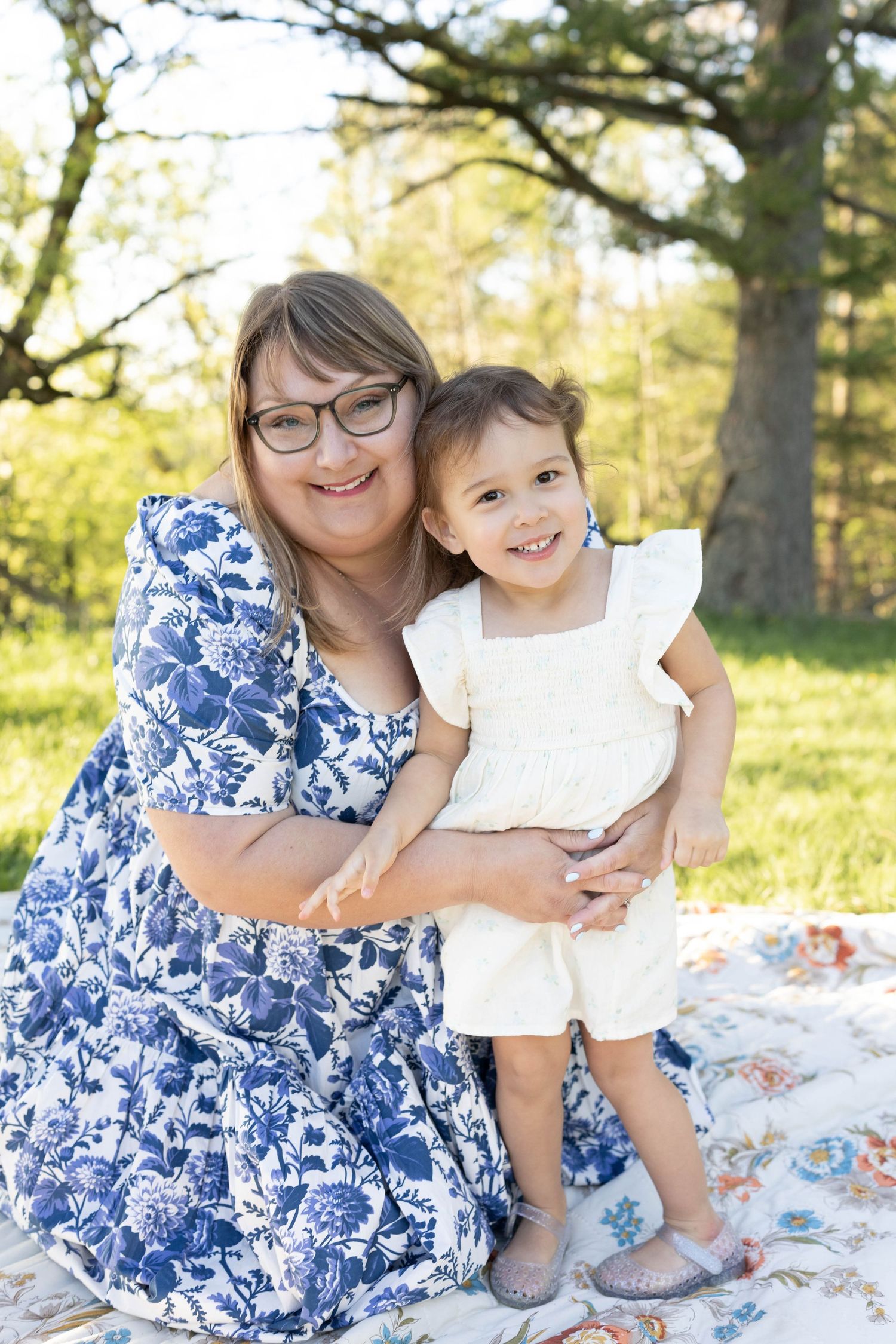 A woman in a blue floral dress sits on a blanket with a small child wearing white on a sunny day outdoors.