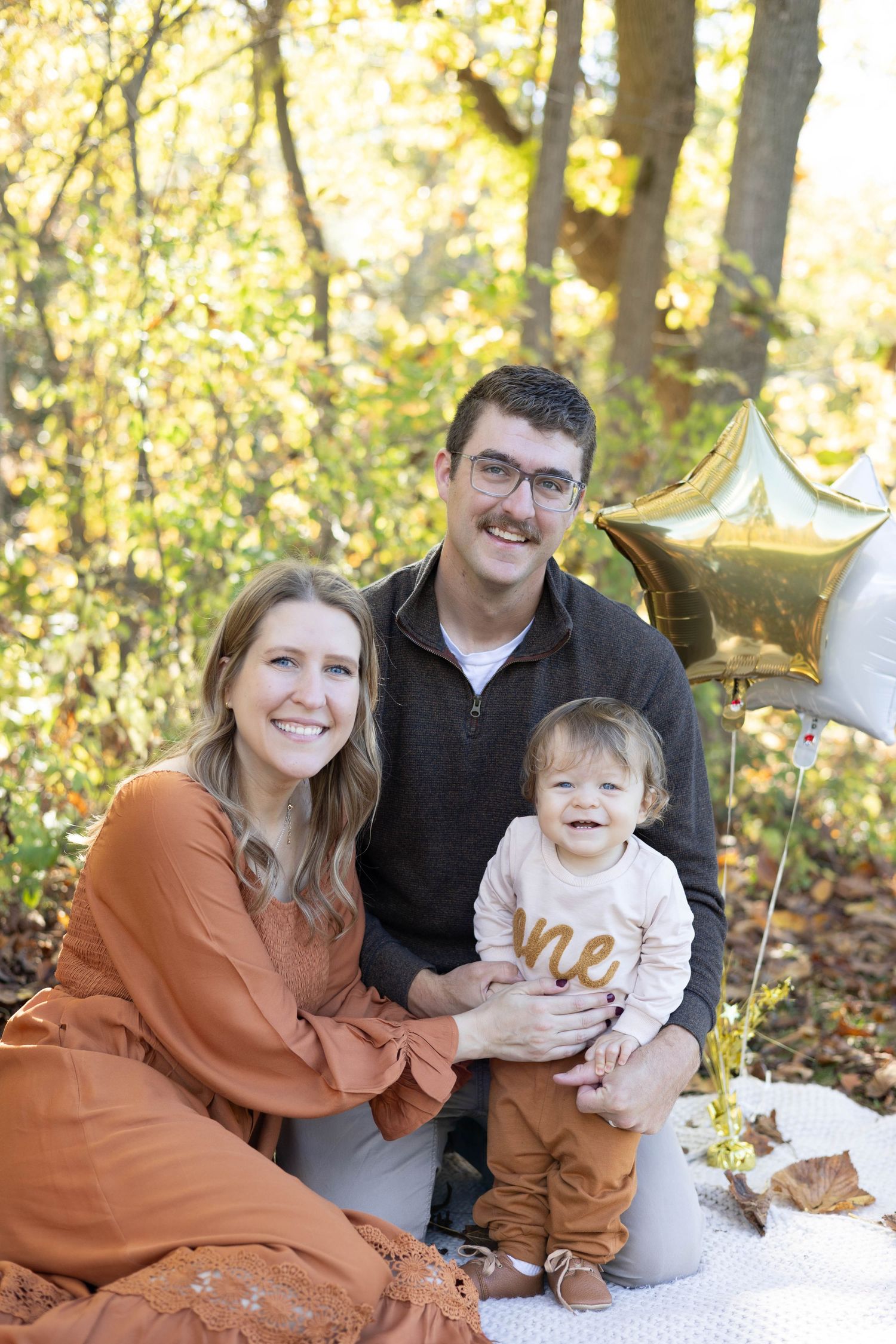 Family photo session in autumn forest with white blanket and gold decorations for baby's first birthday celebration.