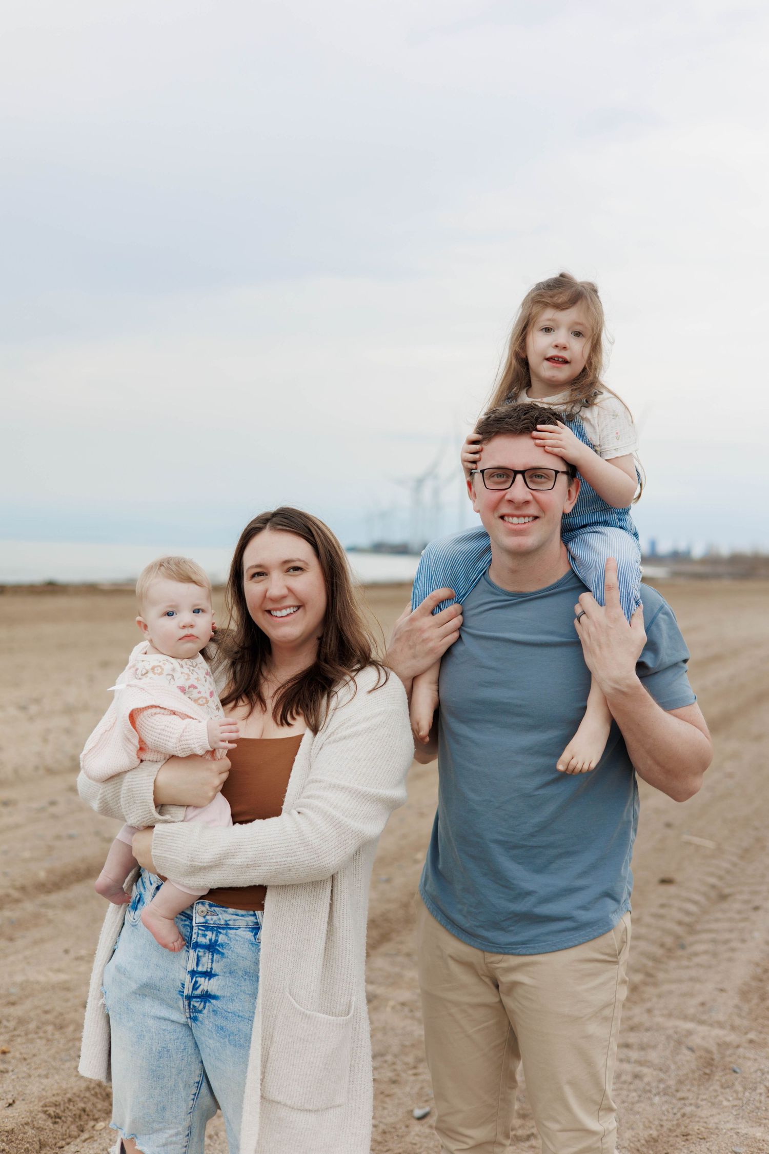 Happy family of four posing on sandy beach, parents holding two young daughters on cloudy day.