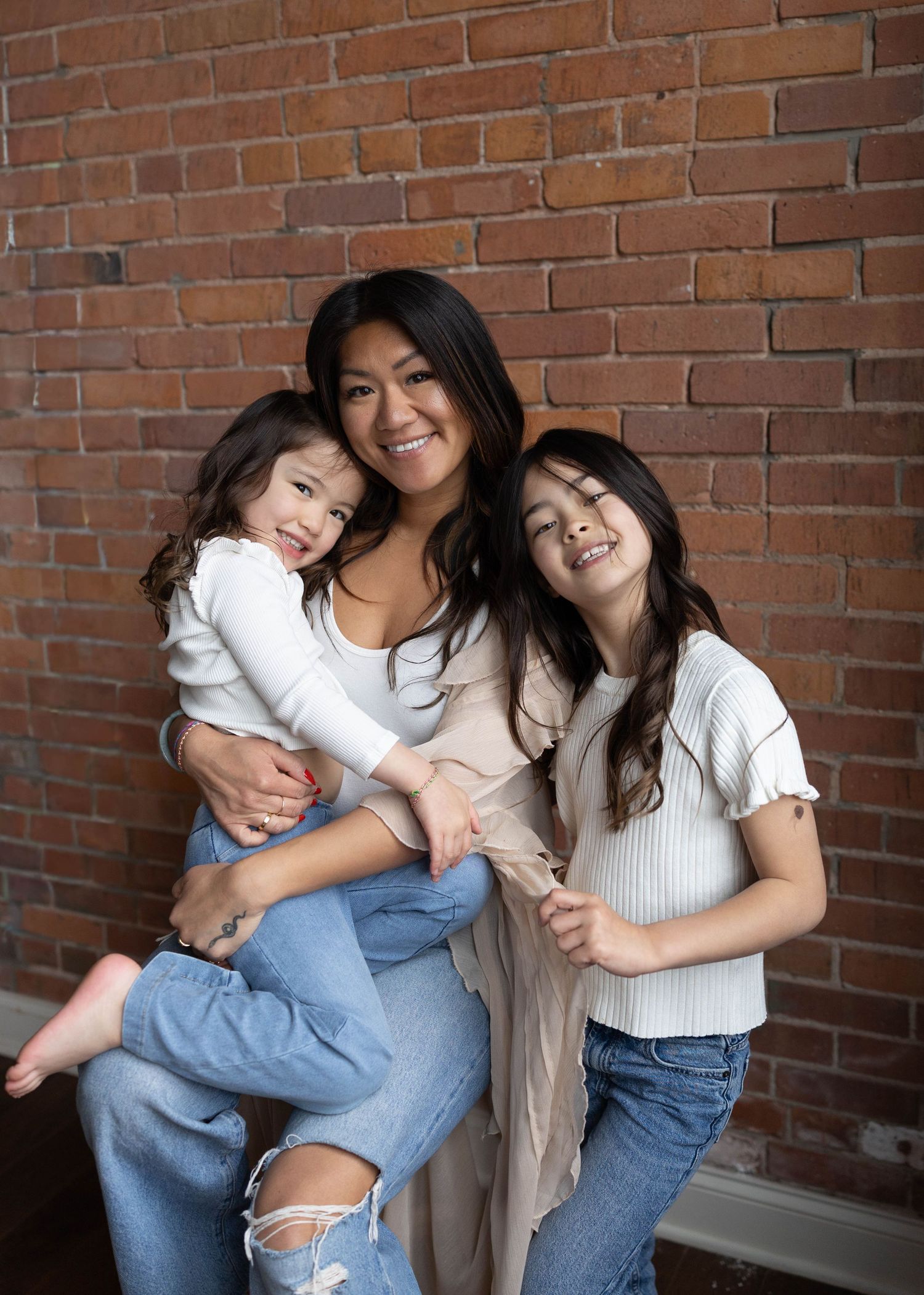 A smiling woman holds a toddler while a young girl leans in for a warm family portrait against a brick wall.