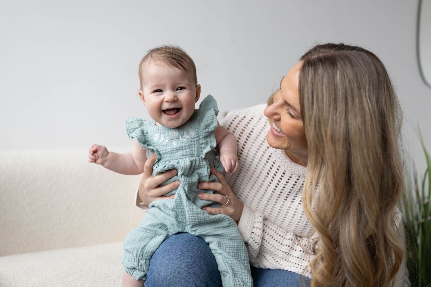 A joyful baby in a teal ruffle outfit laughs while a smiling young mother holds her on a white couch.