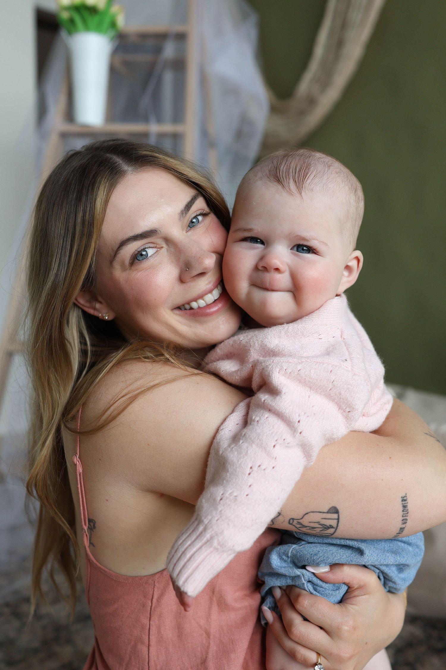 Smiling young mother with long blonde hair holds her happy baby in a pink outfit, sharing a joyful embrace indoors.