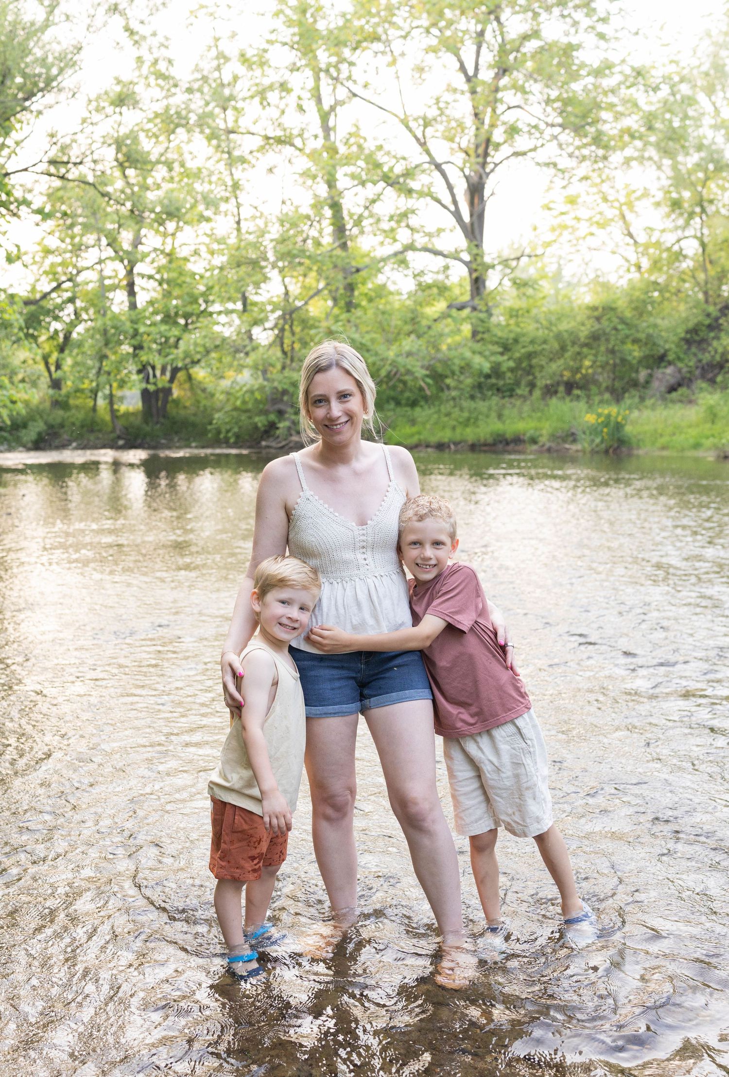 Three people stand together in a shallow creek on a summer day with lush trees in the background.