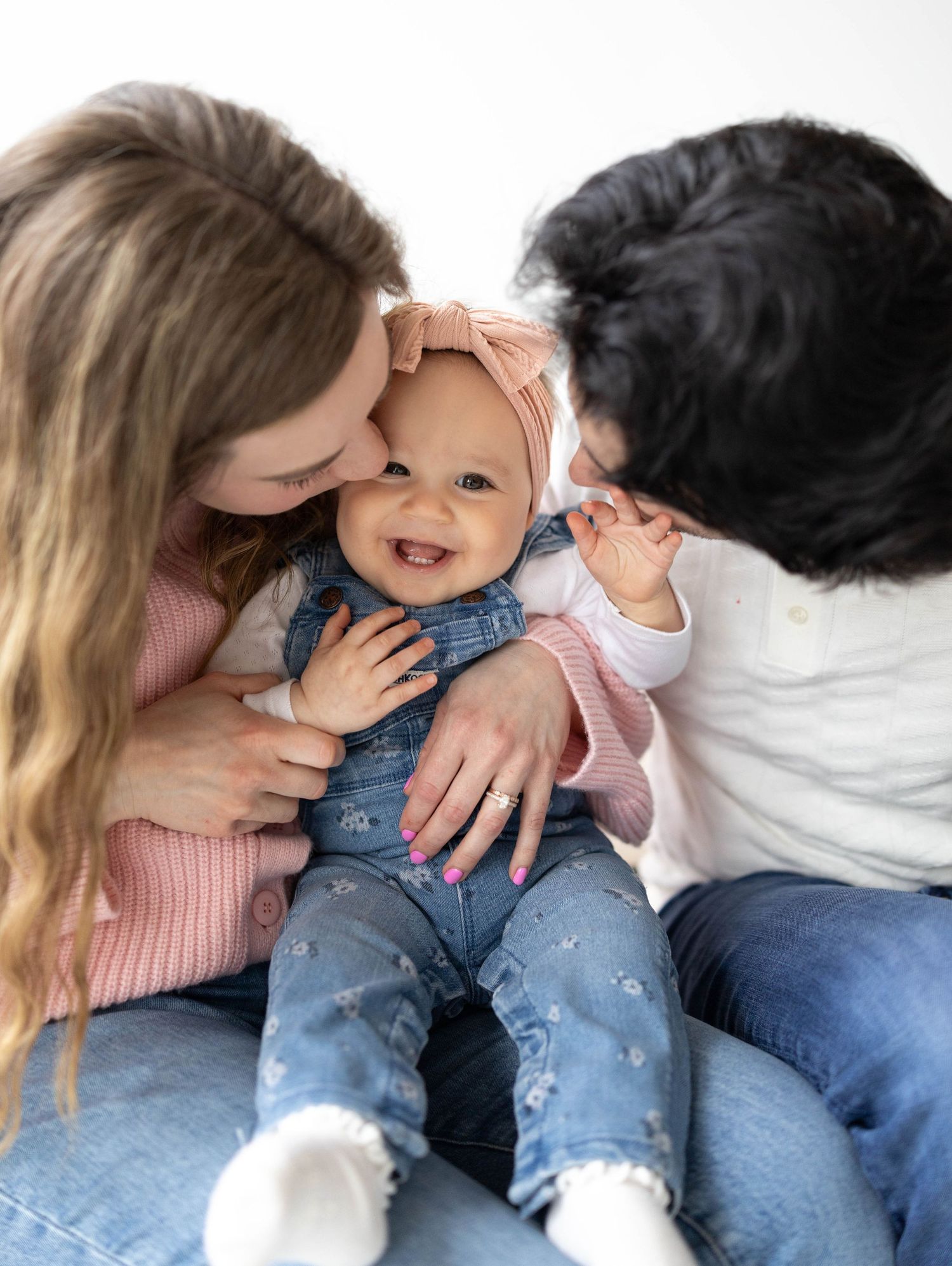 A baby wearing denim overalls and a headband smiles brightly while being embraced and shown affection.
