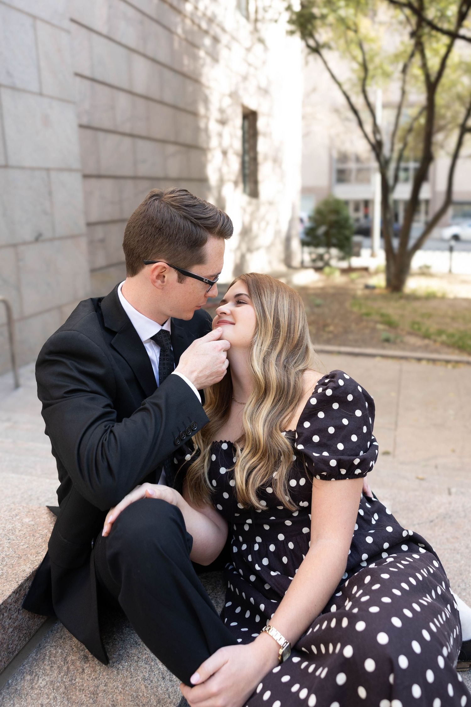 Romantic couple in vintage-inspired attire share an intimate moment on stone steps, she in polka dot dress, he in suit.