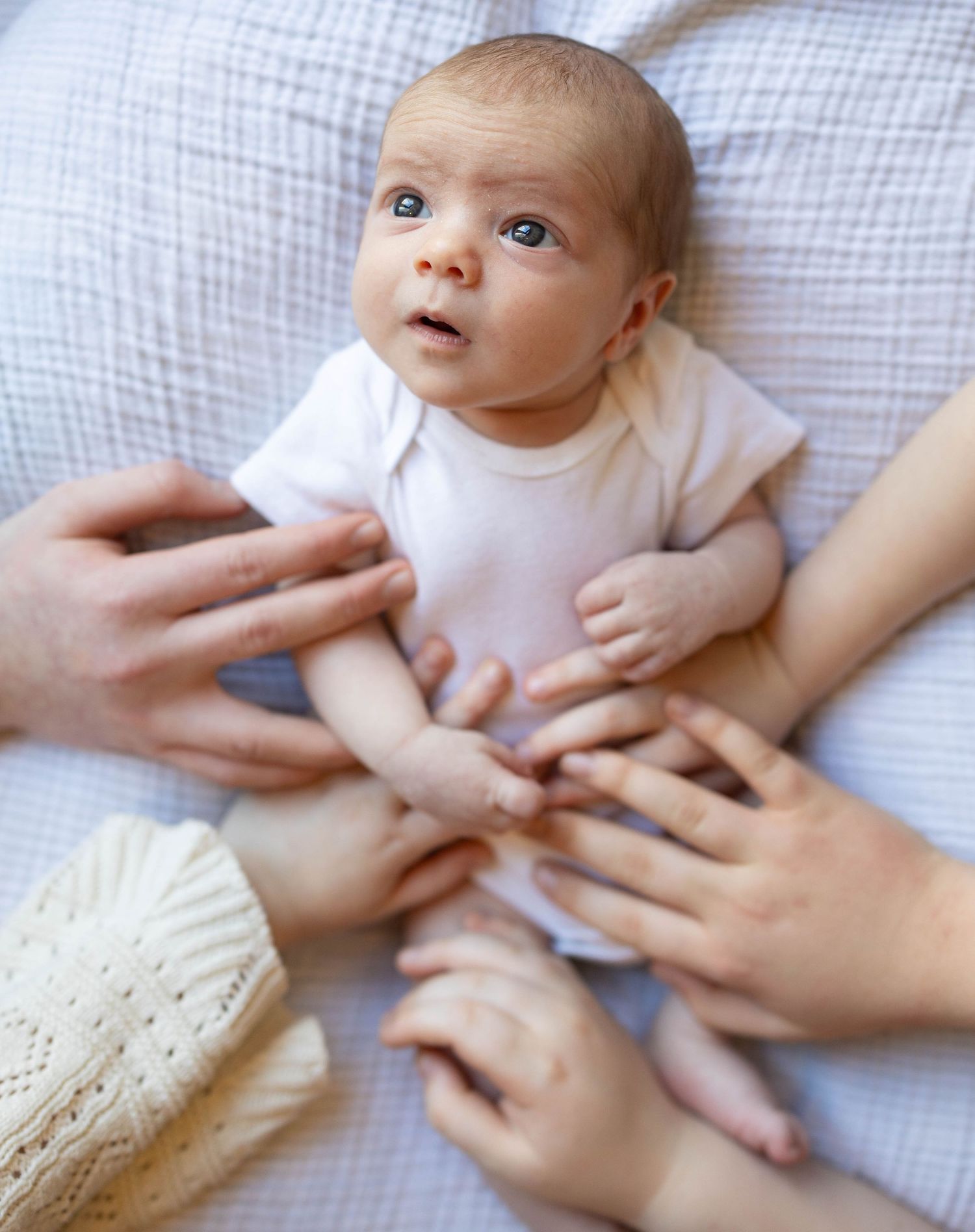 A baby in a white onesie lies on light colored bedding while gentle hands reach down to provide comfort and care.