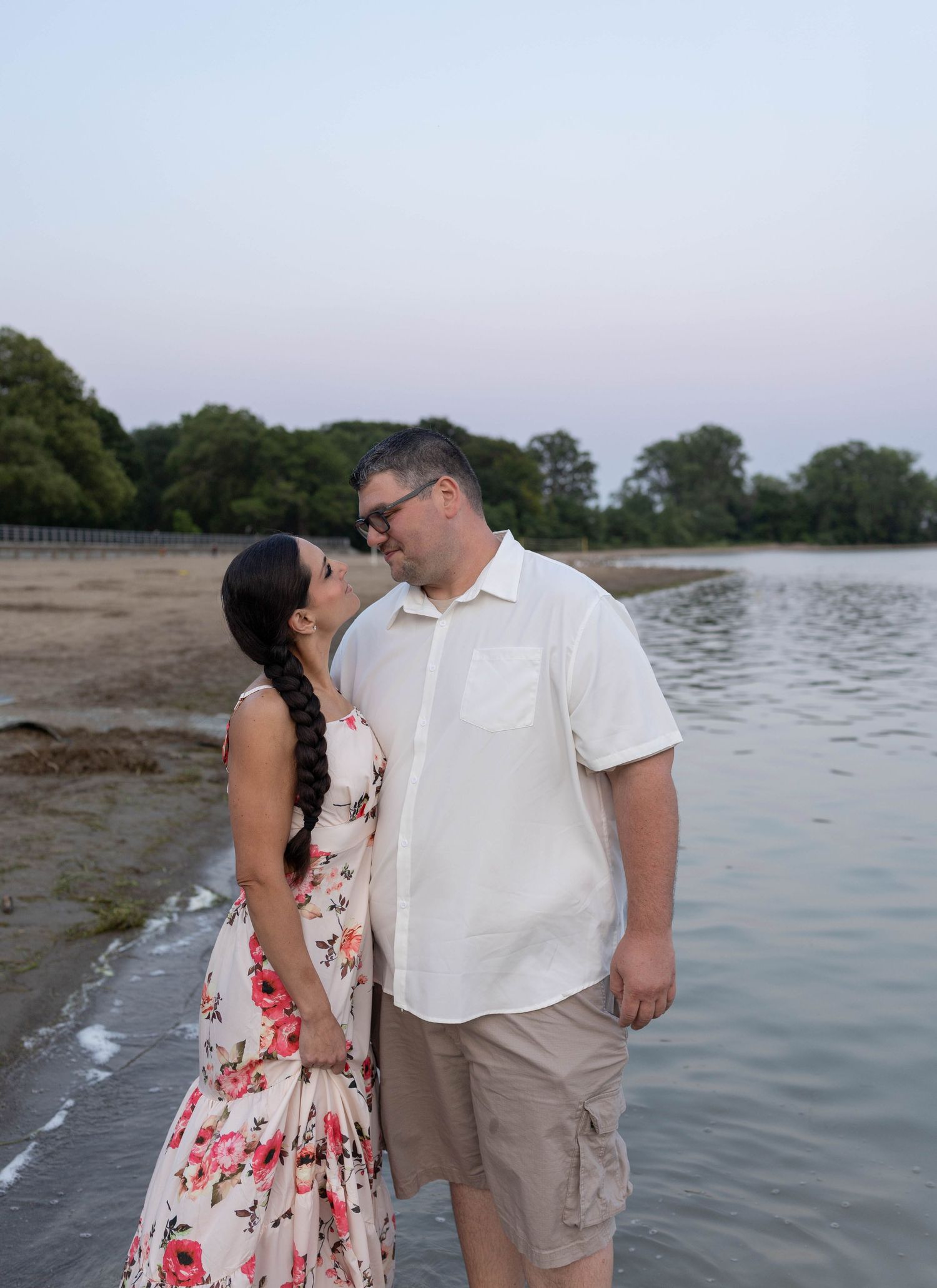 Couple sharing a romantic moment by the water's edge at dusk, woman in floral dress, man in white shirt.