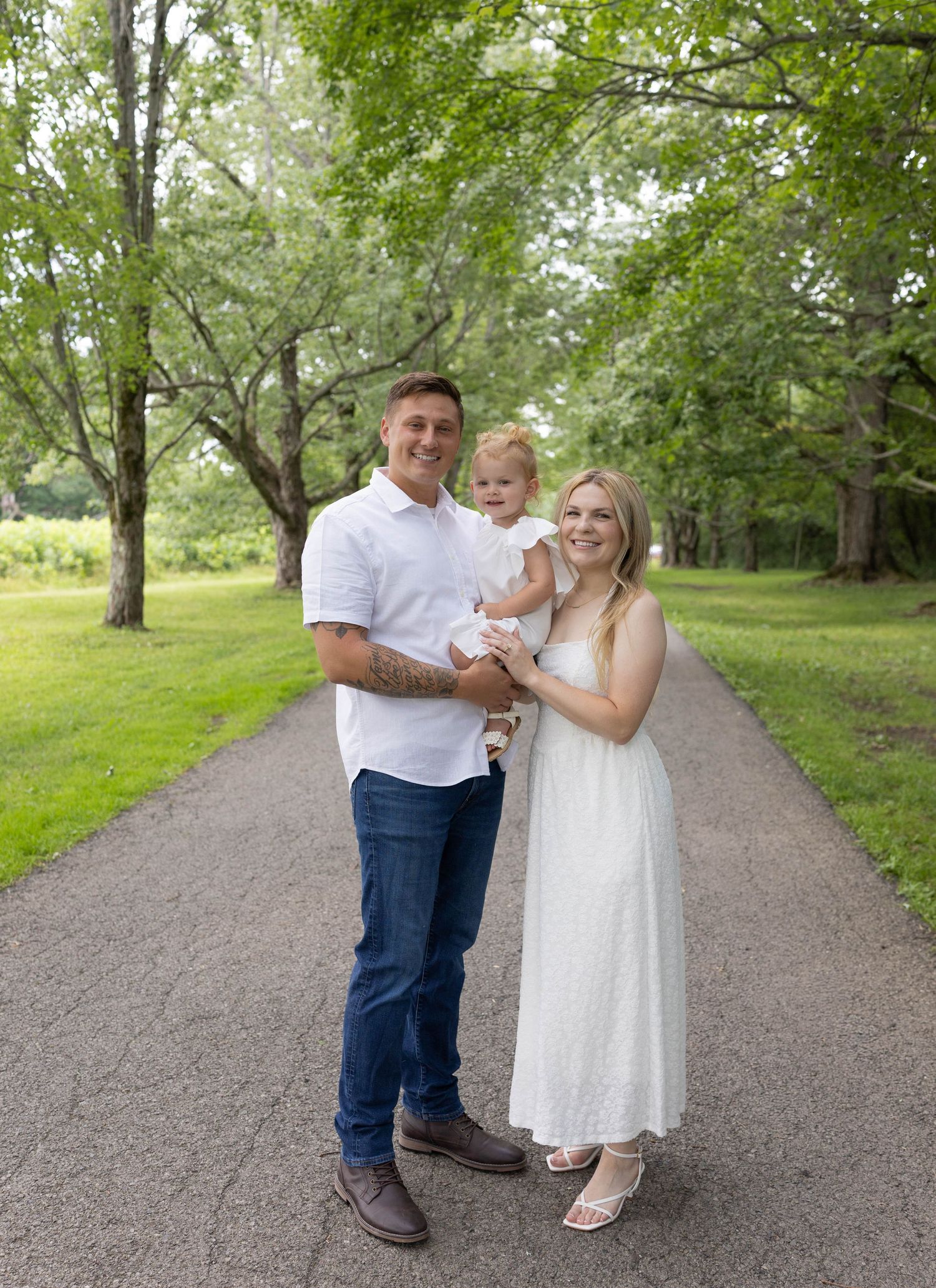A young family with a tattooed man in white shirt, blonde woman in white dress, and baby pose on a tree-lined path.