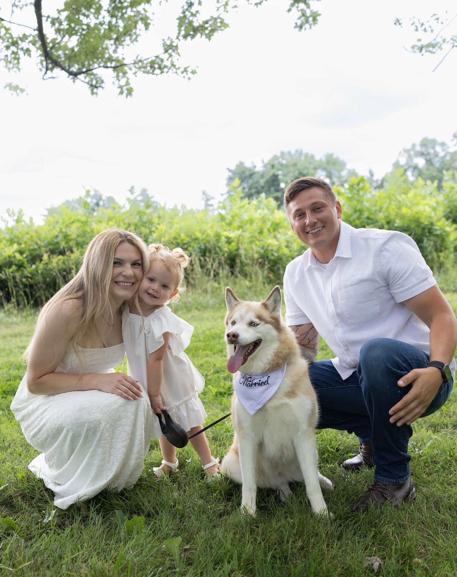 Family posing outdoors on grass with a Husky dog wearing a bandana, surrounded by lush green trees.