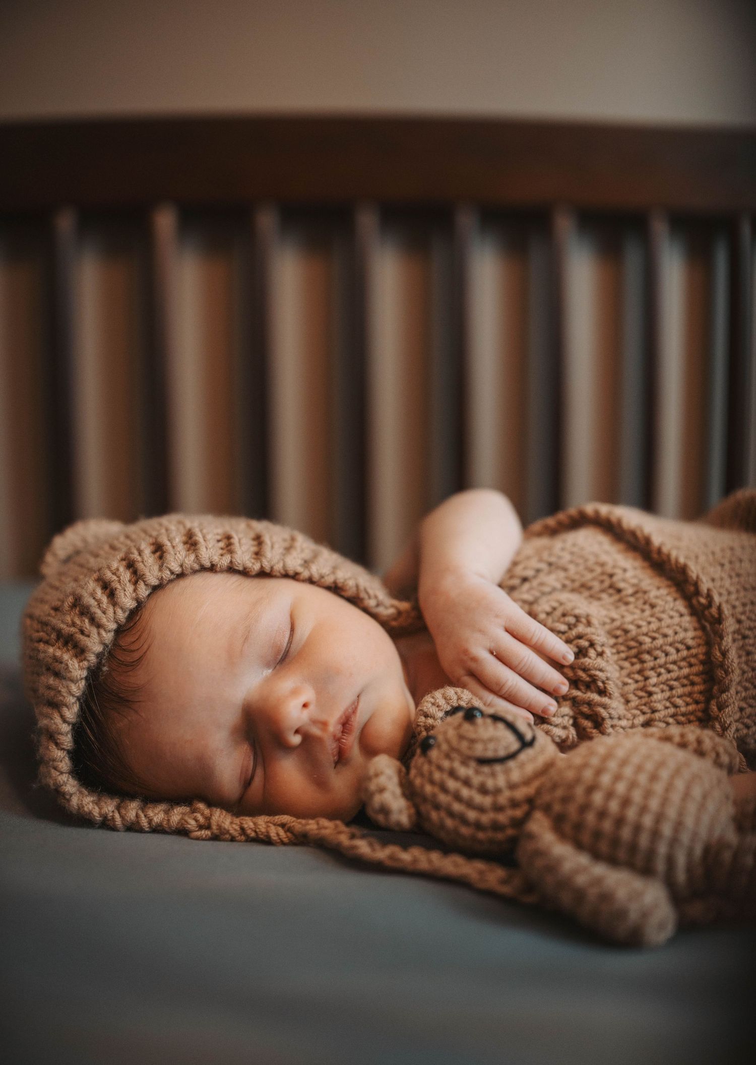 Sleeping newborn baby in brown knit bear outfit holding a crochet teddy bear in a wooden crib.