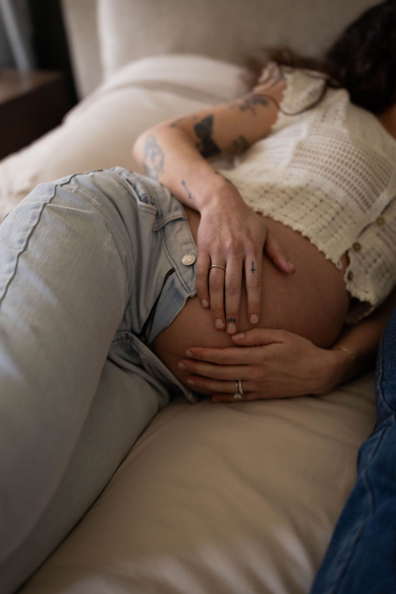 Person wearing light blue jeans and knit sweater resting on white couch with visible tattoos and wedding ring.