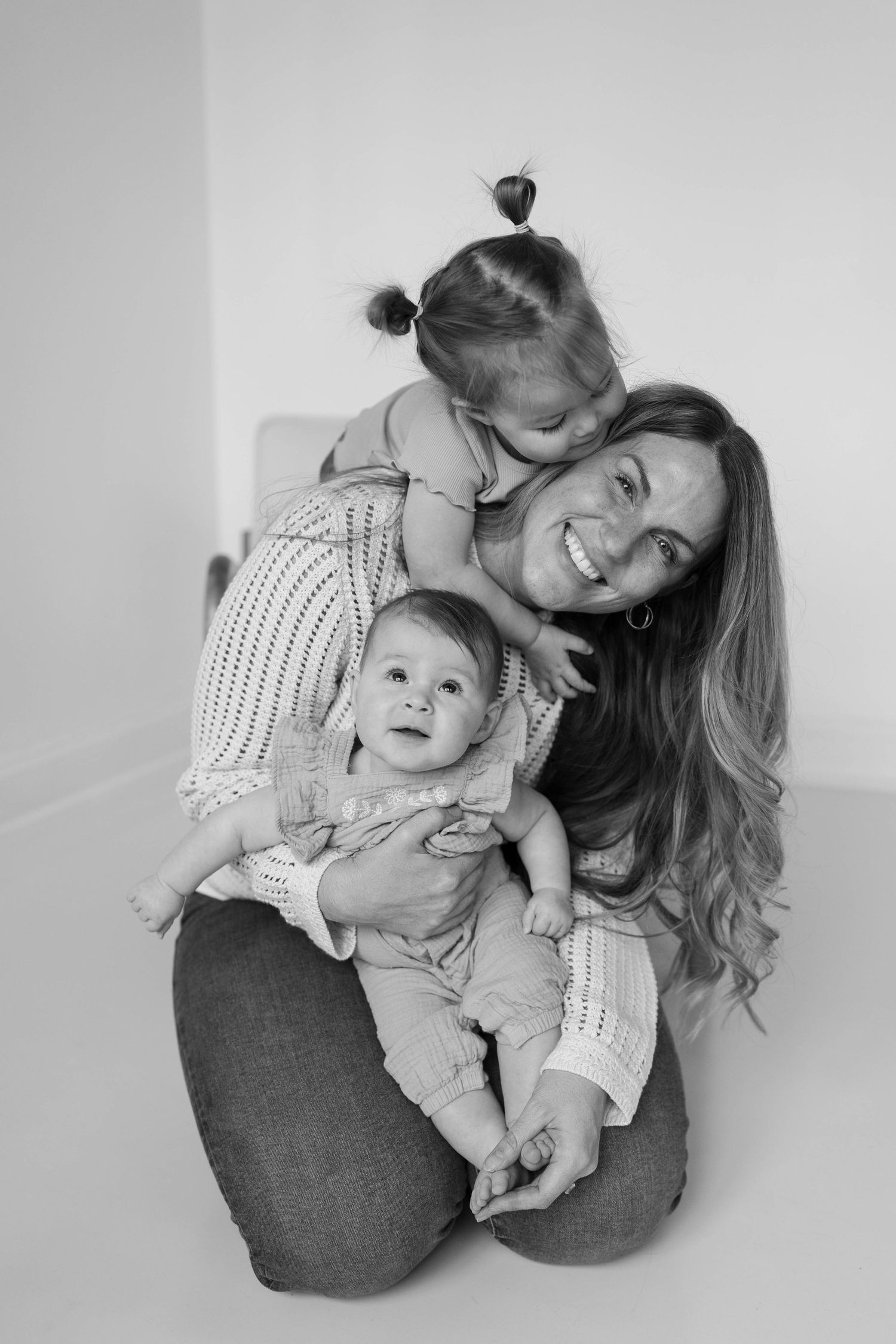 Black and white photo of a smiling mother sitting on floor holding baby while toddler hugs her from behind.