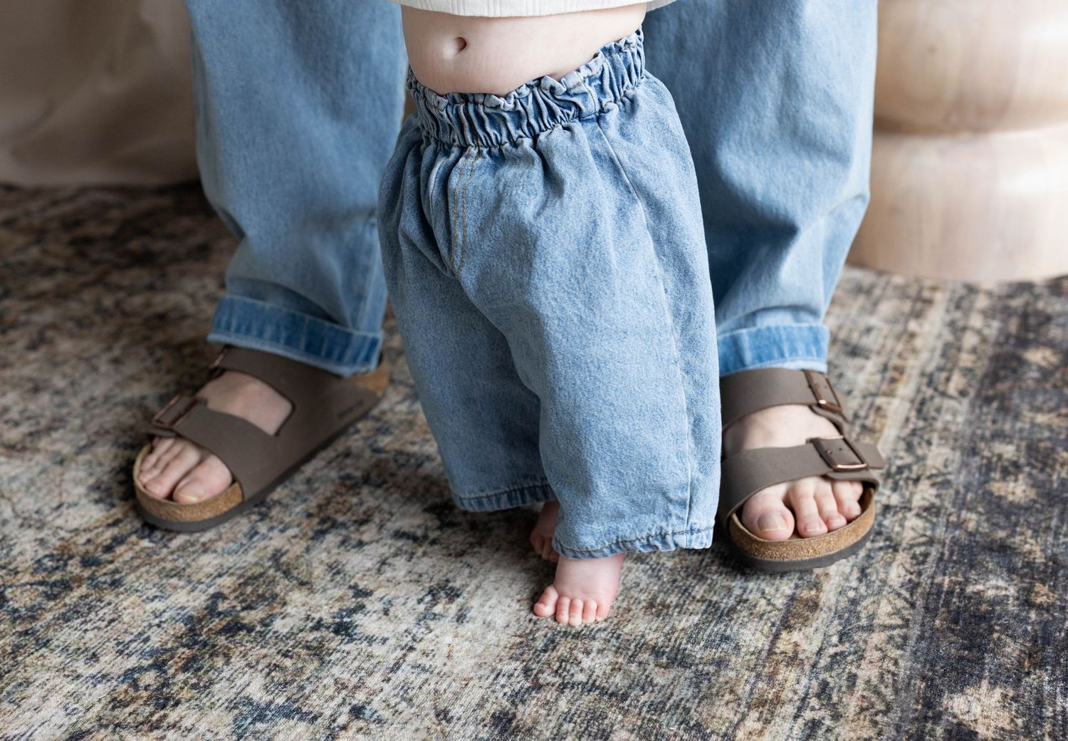 Toddler in blue linen pants standing barefoot beside adult wearing brown Birkenstock sandals on a patterned rug.
