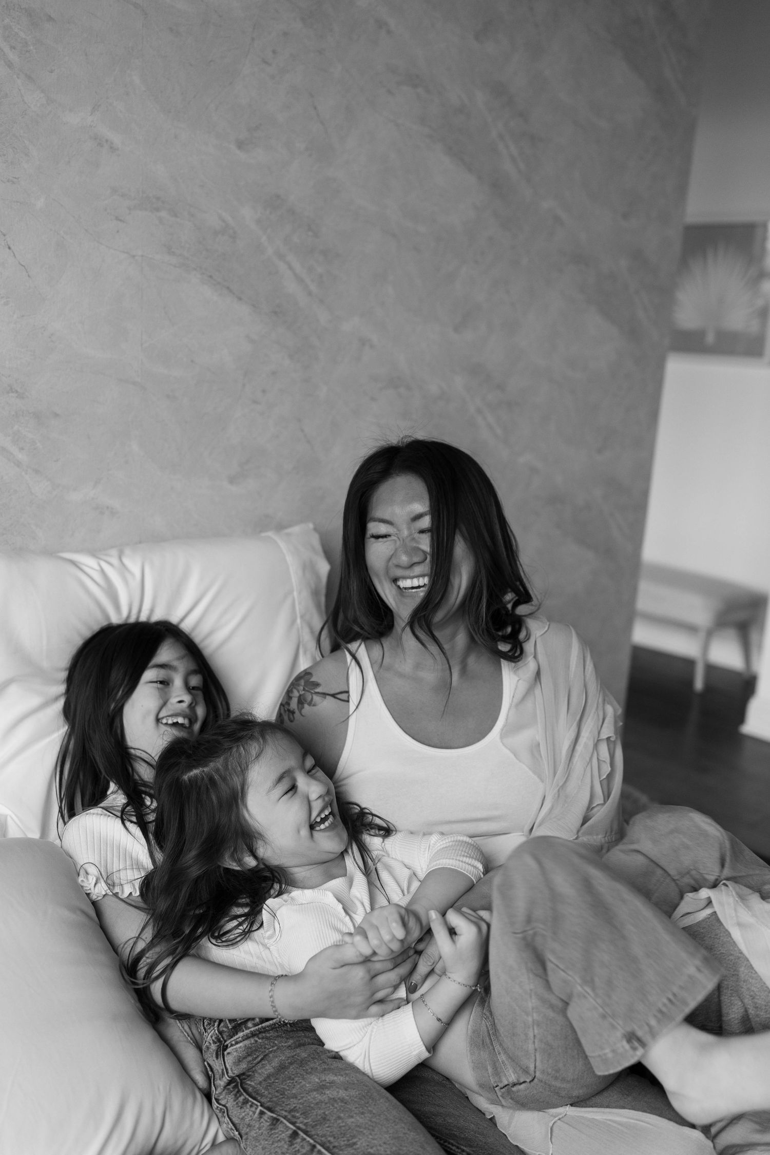 Black and white photo of a mother laughing joyfully with her two young daughters on a cozy sofa at home.