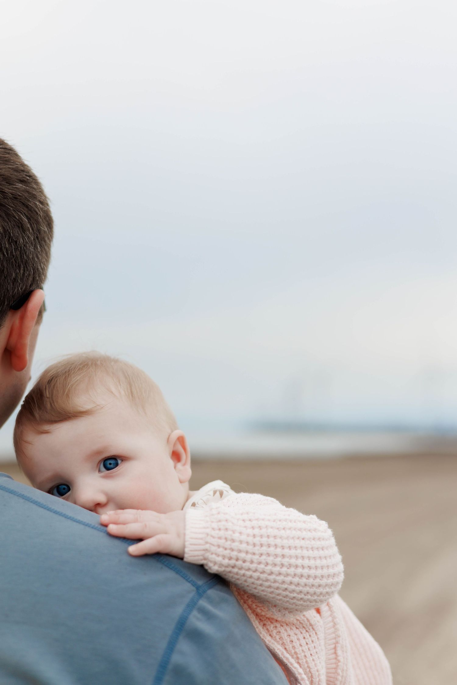A father holds his blue-eyed baby on a beach, the infant in a pink knit outfit looking over his shoulder.