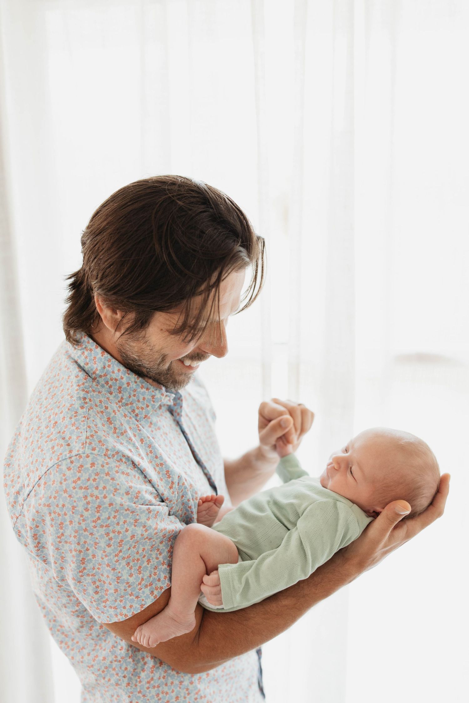 A parent in a patterned shirt holds and plays with a small baby dressed in light green clothing.