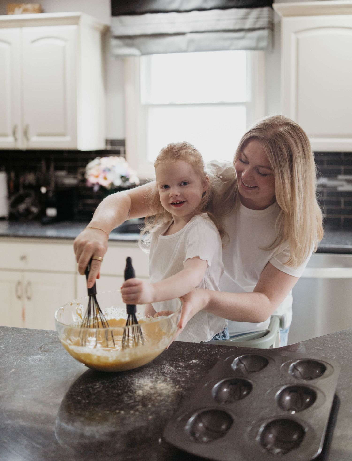 A parent and small child whisk ingredients together in a mixing bowl while baking in a white kitchen.