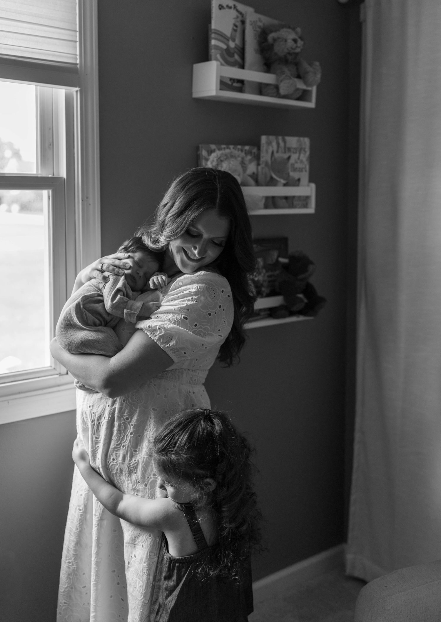 Black and white photo of a smiling mother holding a newborn while a toddler hugs her in a nursery.