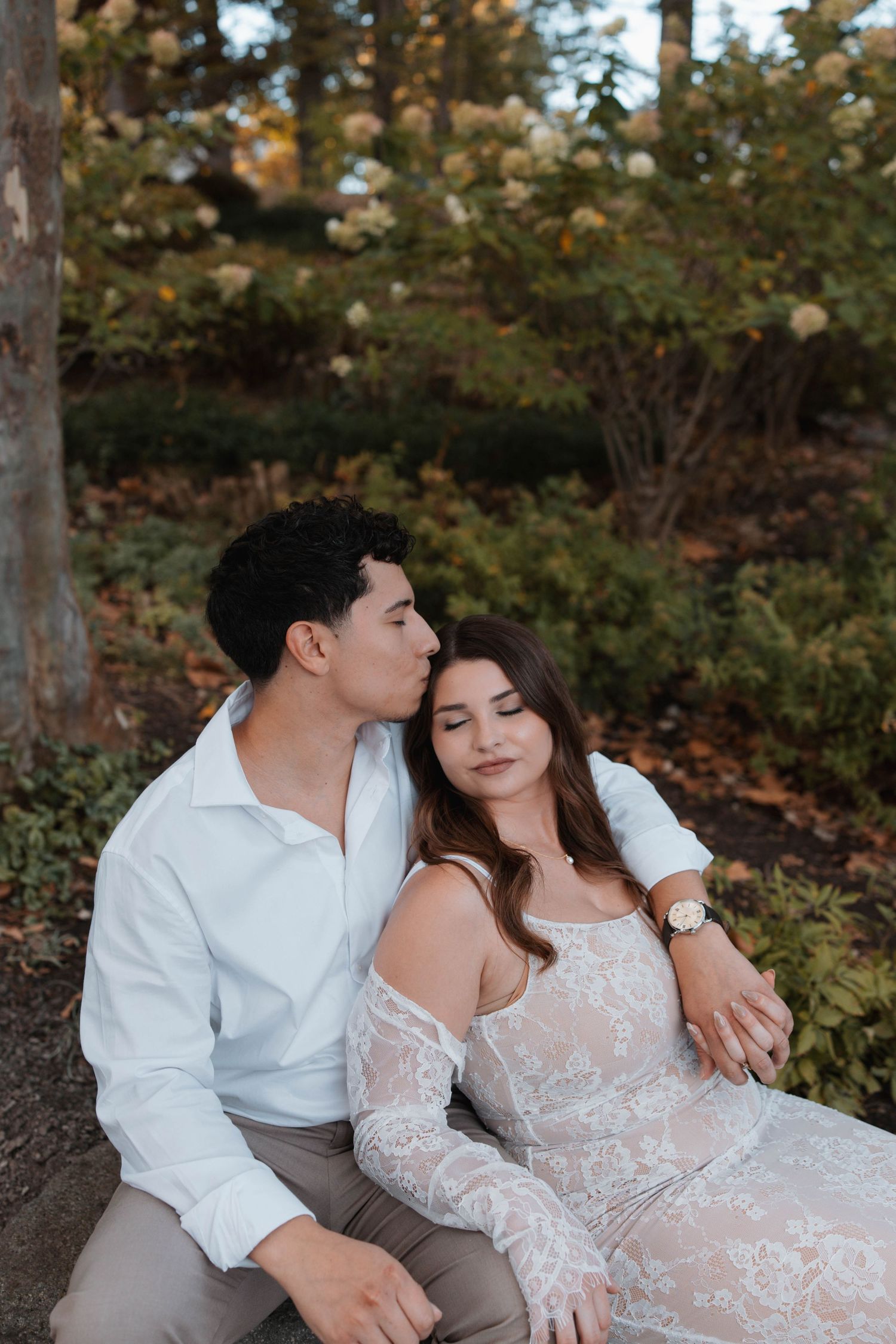 A romantic couple in white attire share an intimate moment outdoors among autumn trees and foliage.