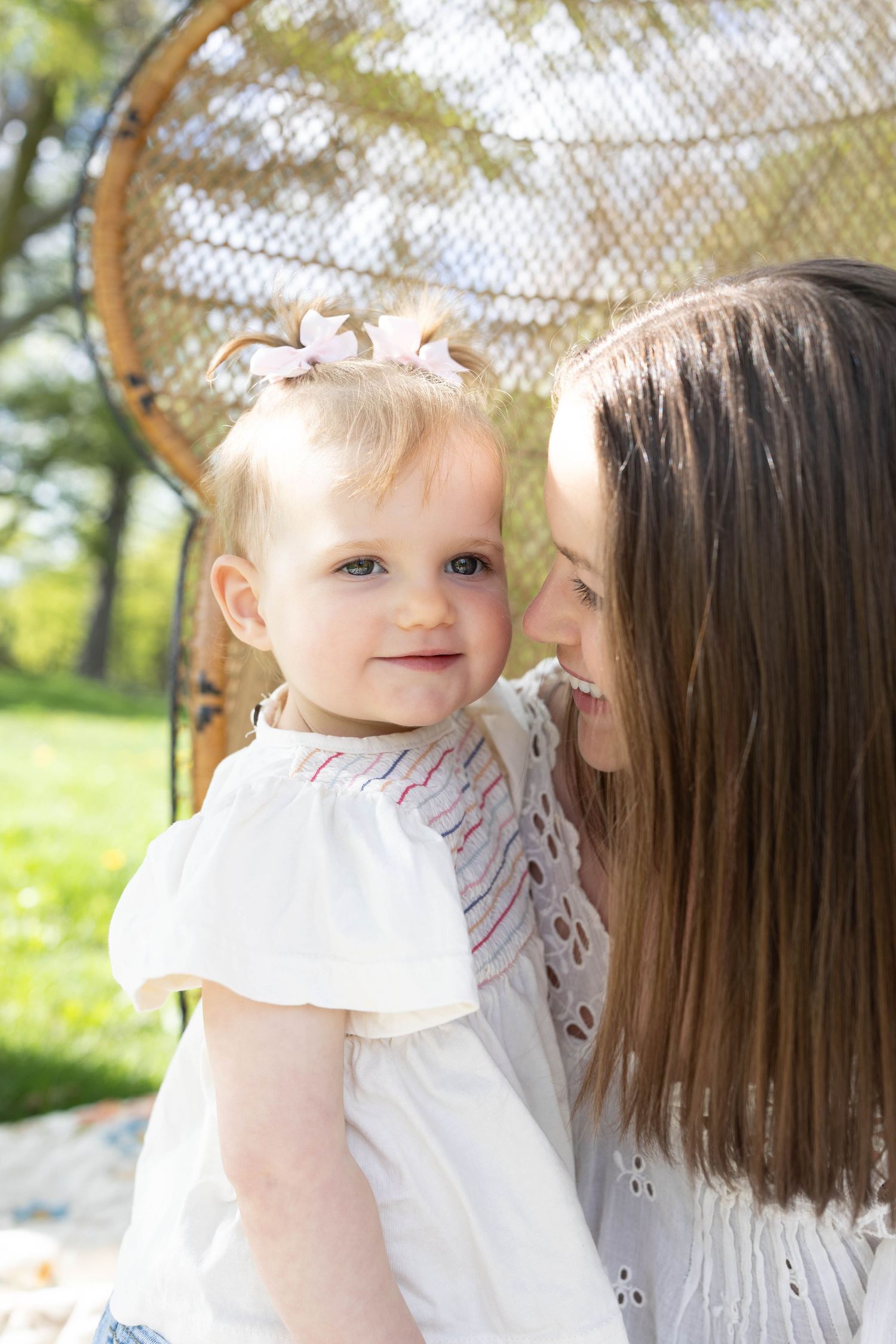 Mother and baby in white dresses share a tender moment outdoors in front of a wicker chair on sunny day.