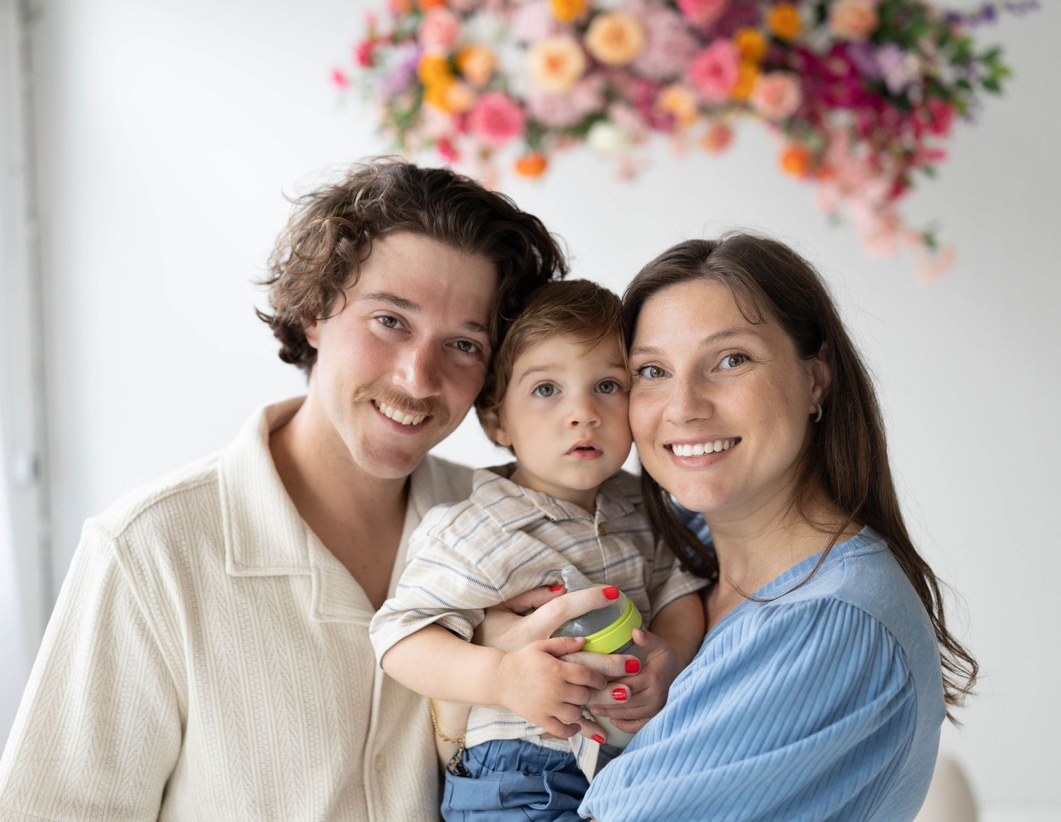A happy family of three poses together in front of a colorful floral backdrop while wearing casual clothing.