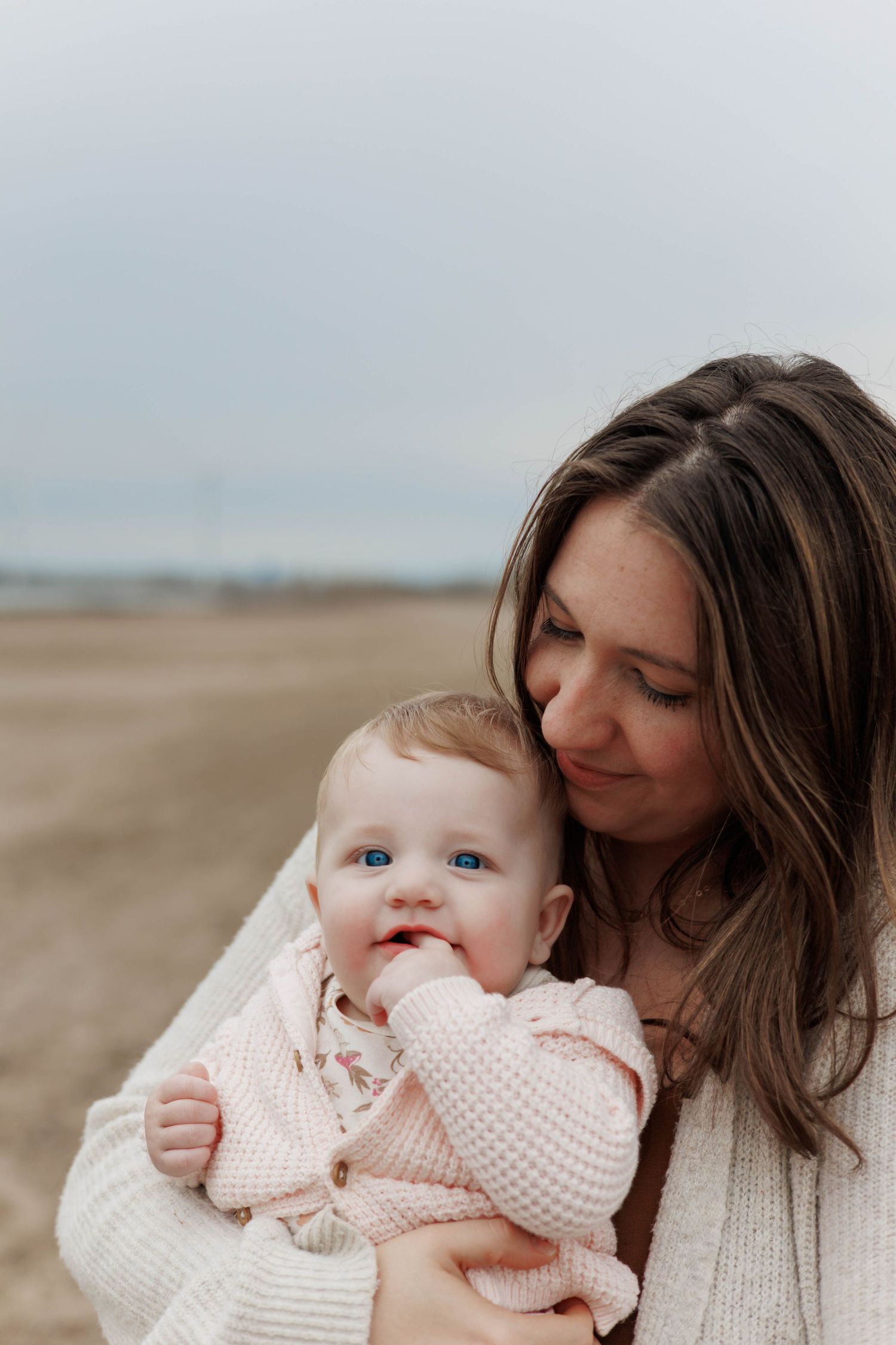 A mother tenderly holds her blue-eyed baby on a sandy beach, both wearing soft pink and white outfits.