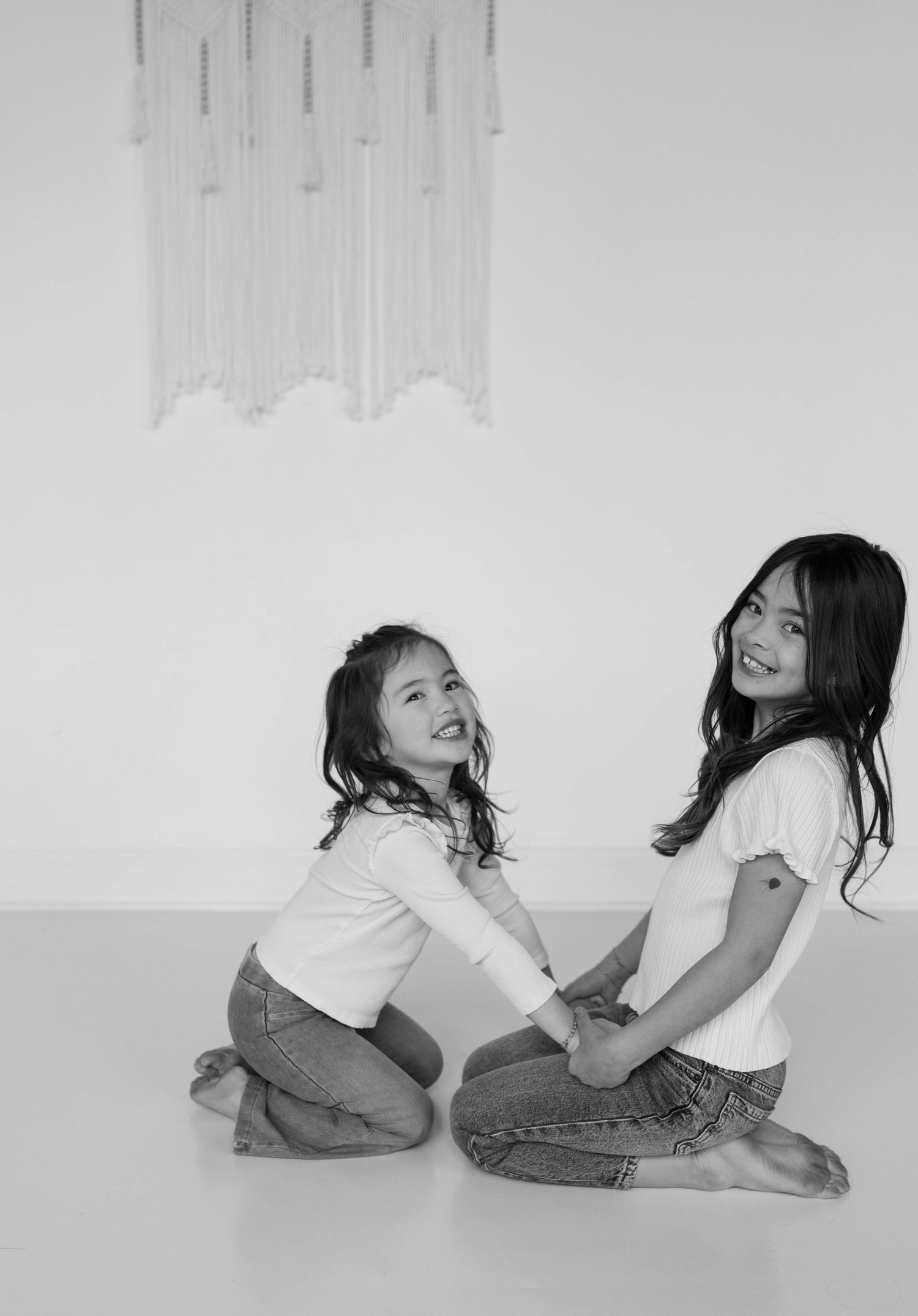 Two young girls in white tops and jeans sit on floor smiling, black and white photo with macrame wall art.