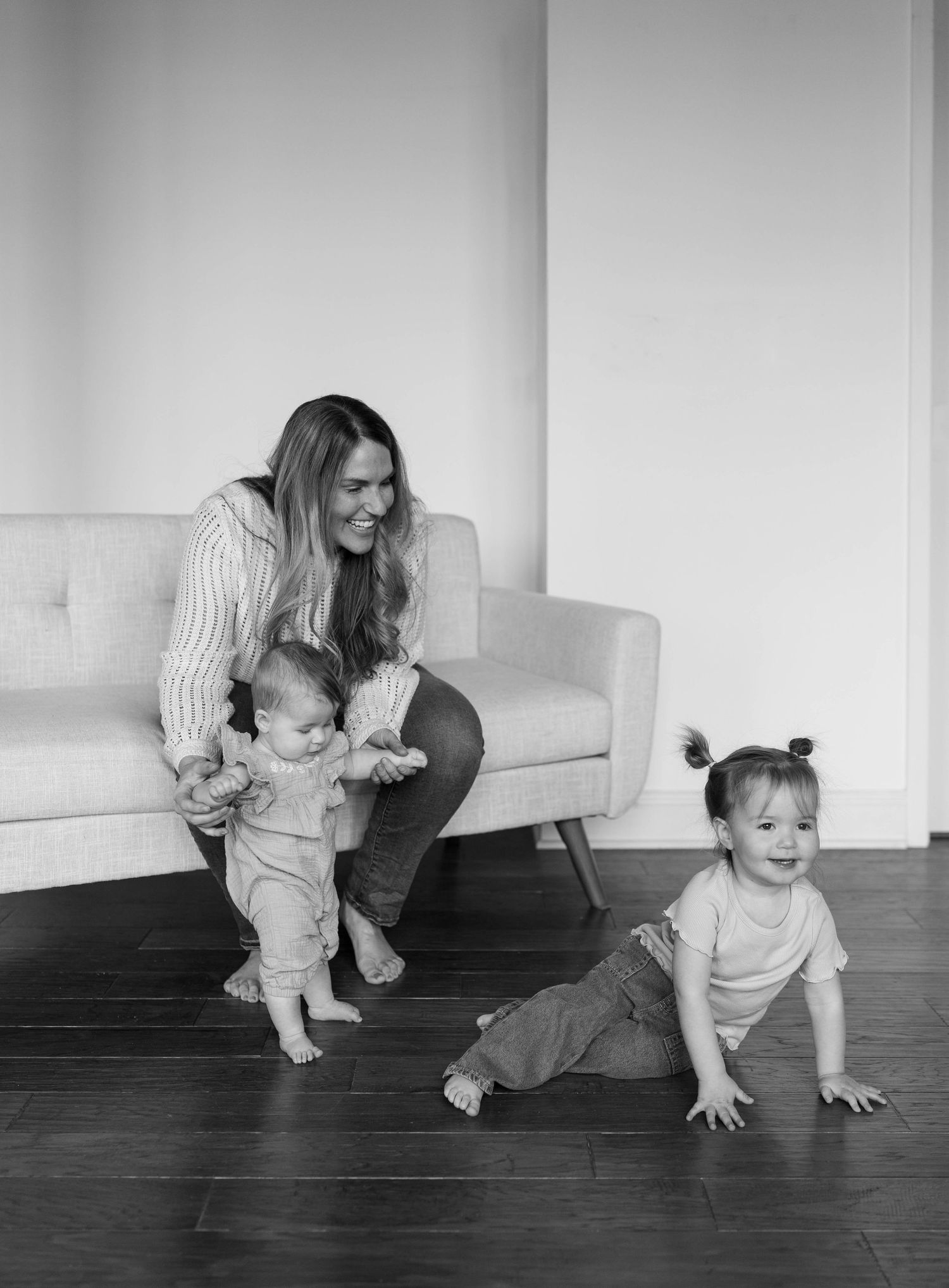 Black and white photo of a smiling woman helping a baby stand near a sofa while a toddler plays on hardwood floor.