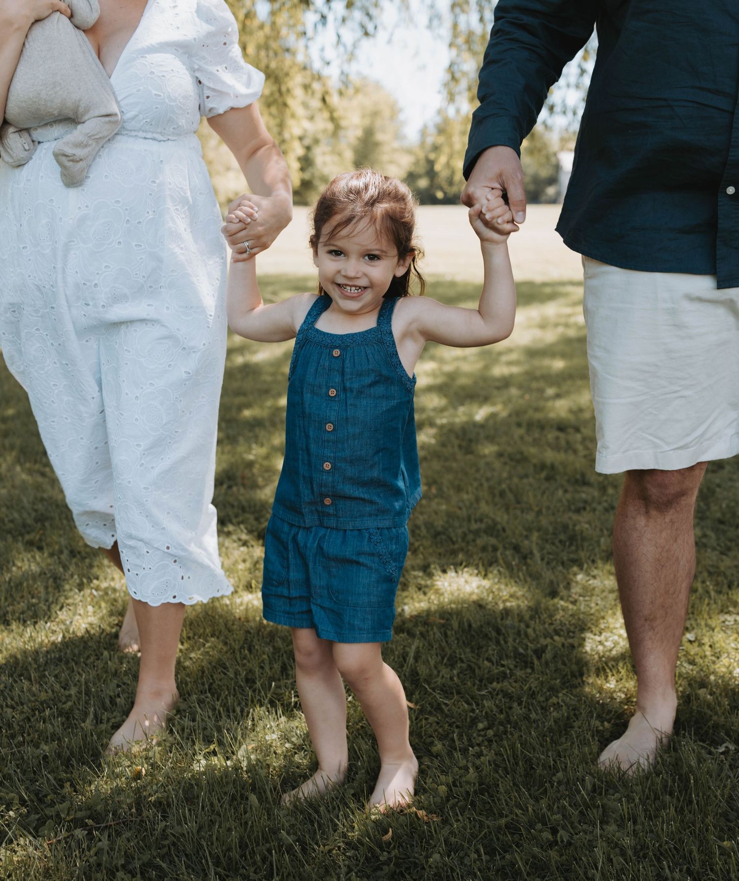 A happy toddler girl in a blue romper holds hands with her parents while standing barefoot on green grass.