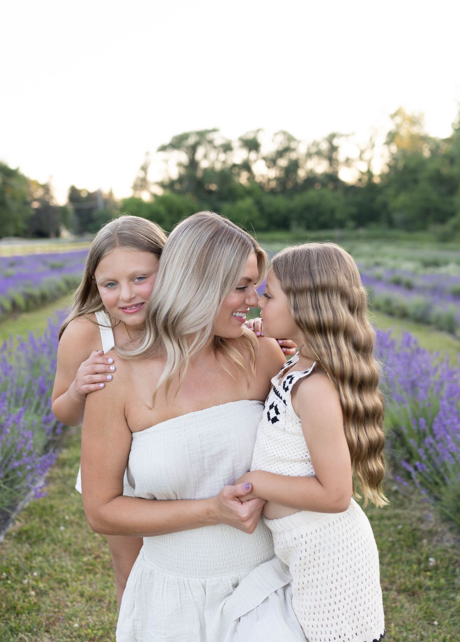Three people share loving embraces in a blooming lavender field at sunset with trees in the background.