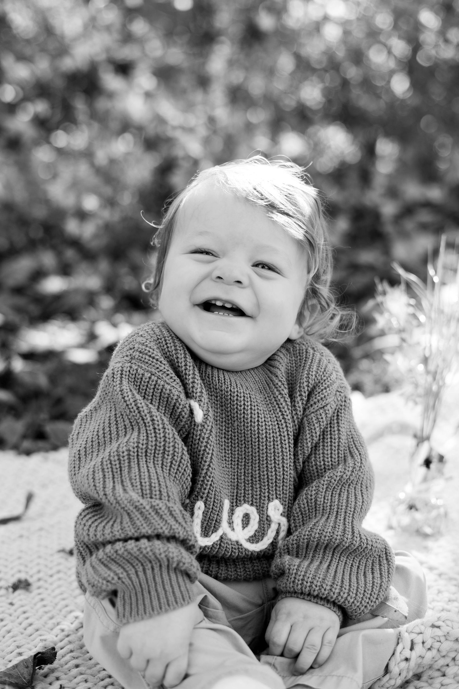 Black and white photo of a happy baby wearing a knit sweater sitting on a blanket outdoors with bokeh background.