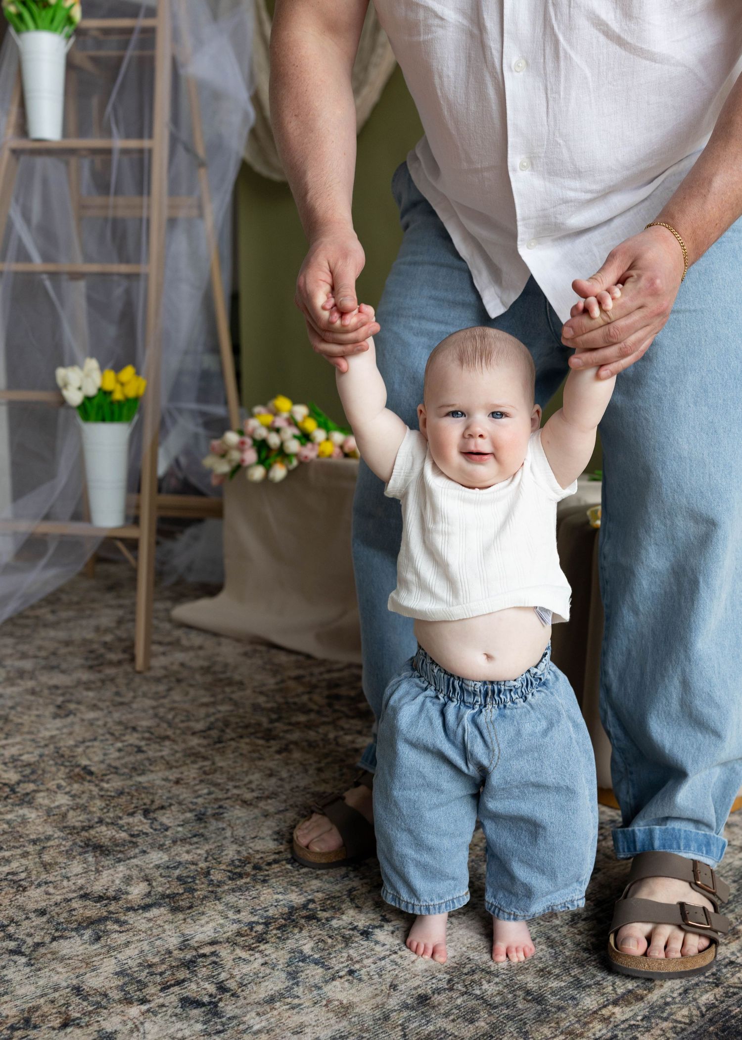 Father helping smiling baby boy take first steps in white onesie and jeans.