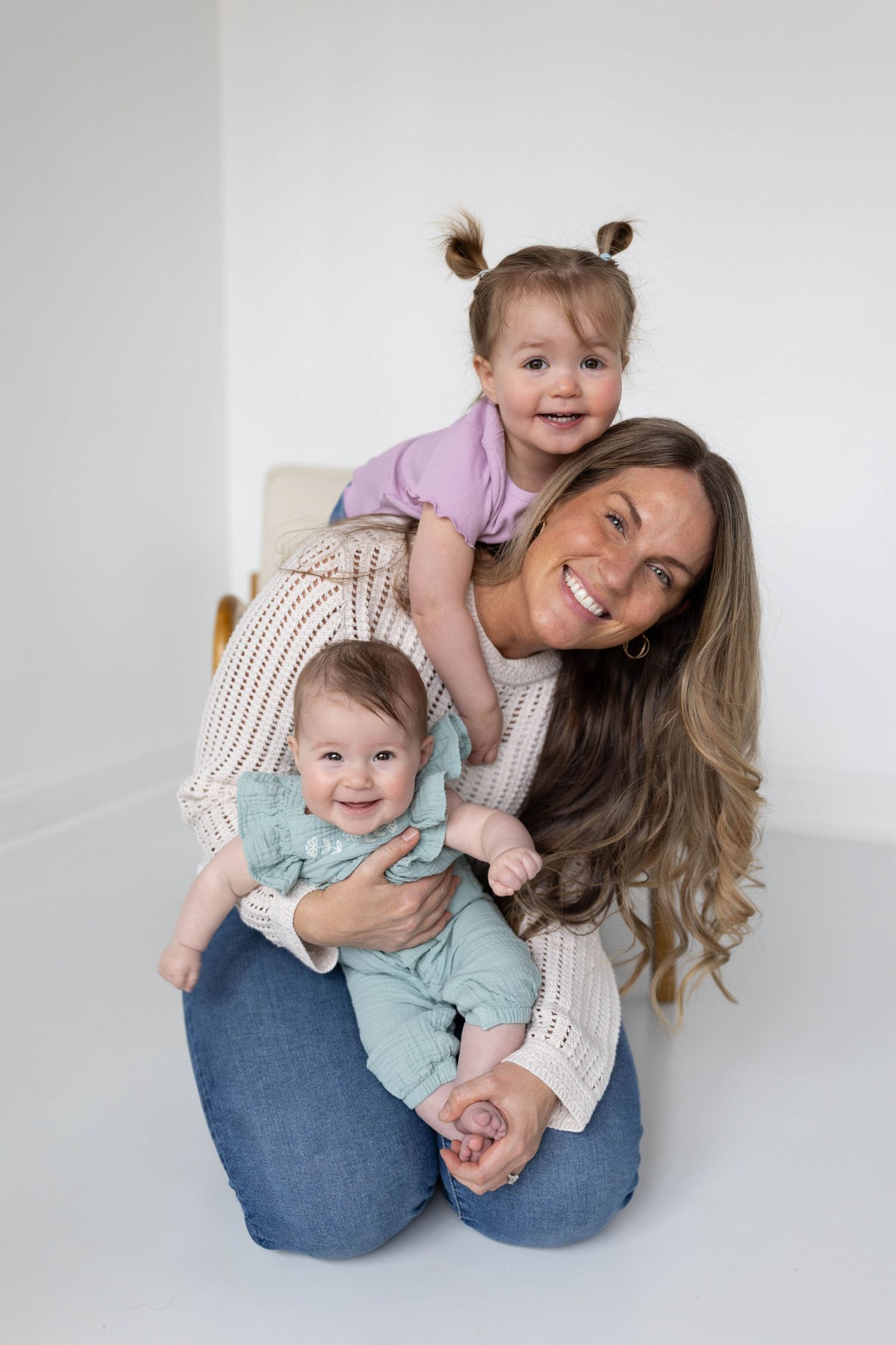 A smiling mother kneels on a white floor holding two young daughters, one baby in teal and a toddler in purple on her back.