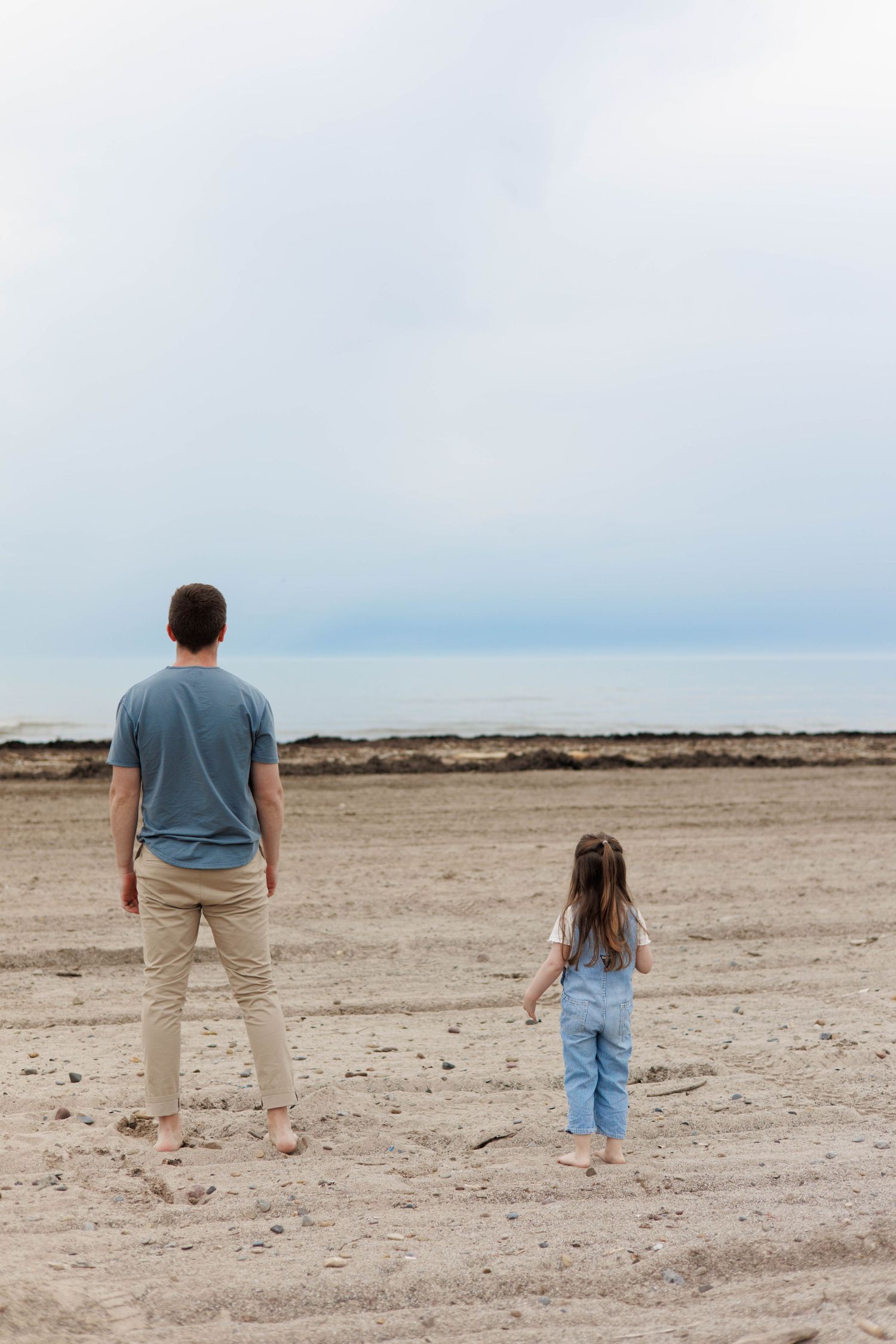 Father and young daughter standing barefoot on sandy beach gazing at ocean horizon together.