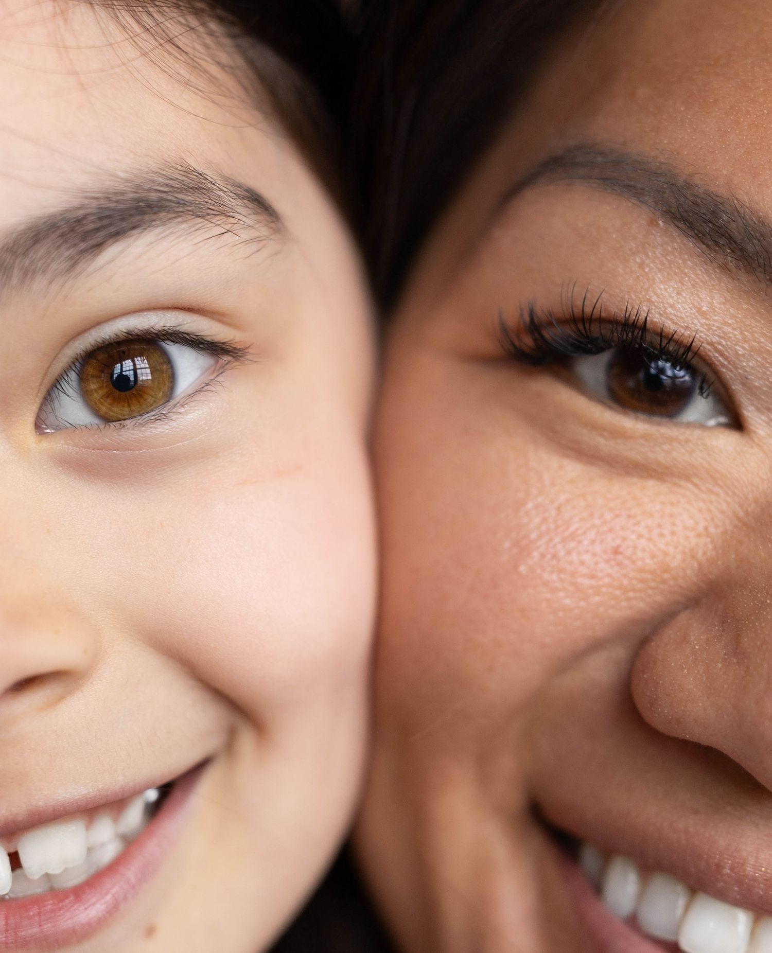 Close-up portrait of two smiling women with glowing skin, showcasing diverse beauty and natural makeup looks.