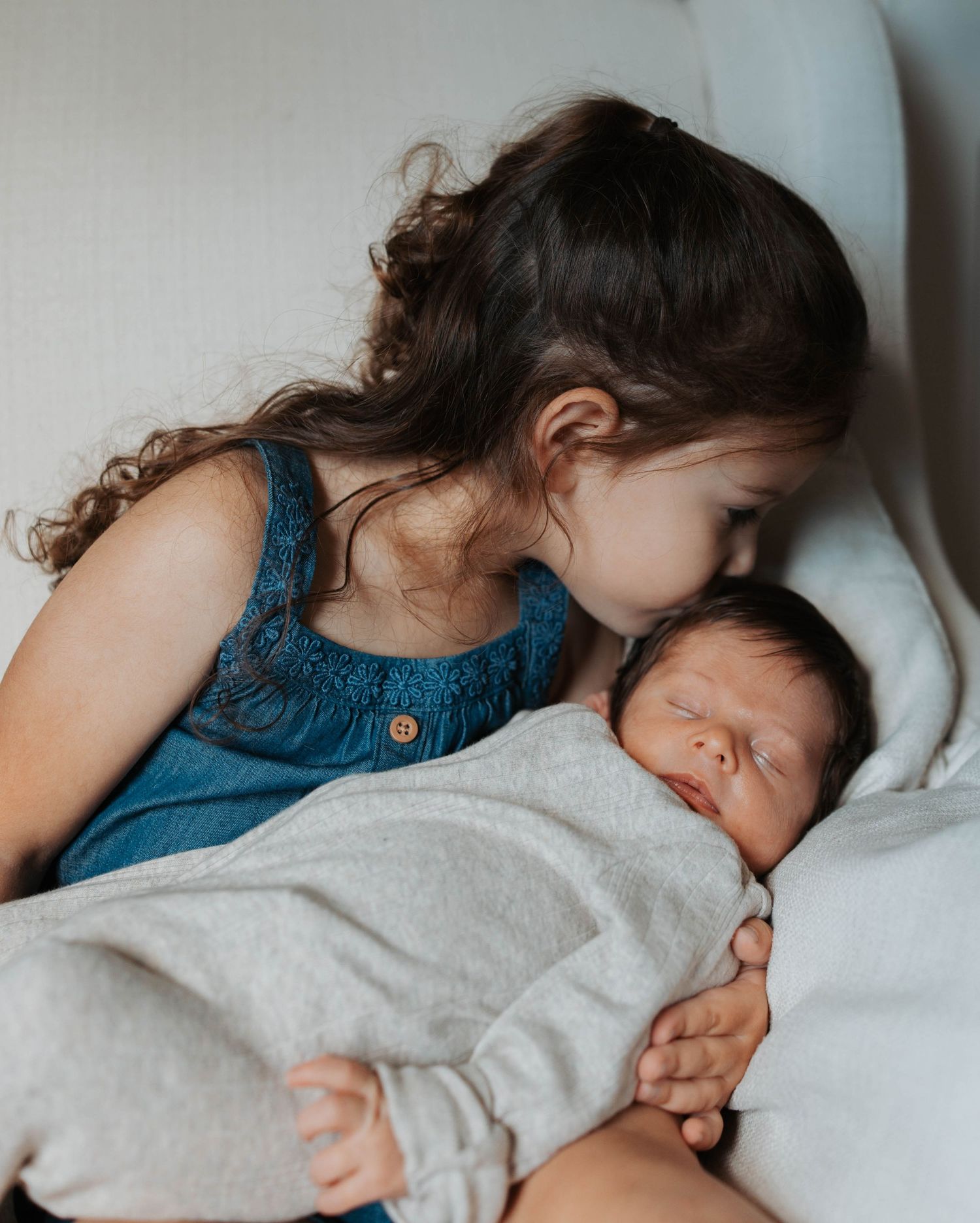 A young girl gently kisses a sleeping newborn baby wrapped in a gray blanket on a white chair.