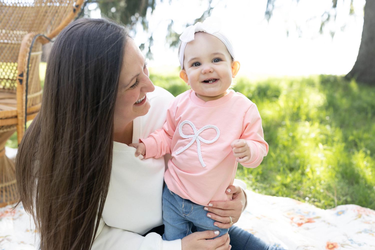 A parent and baby share a happy moment outdoors while sitting on a blanket in a sunny garden setting.