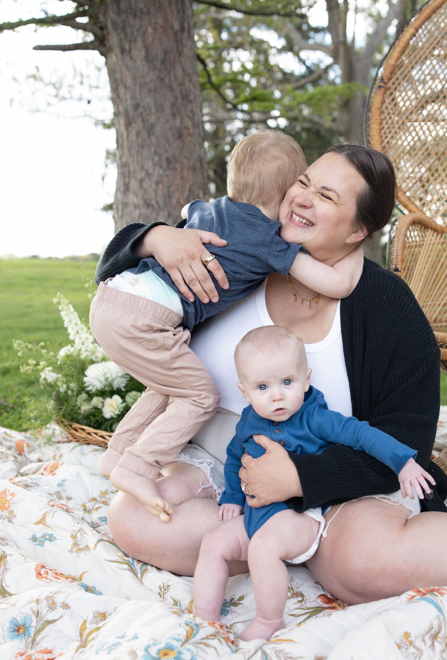 Sweet outdoor family moment captured on a floral blanket with woven chair and pine trees in background.