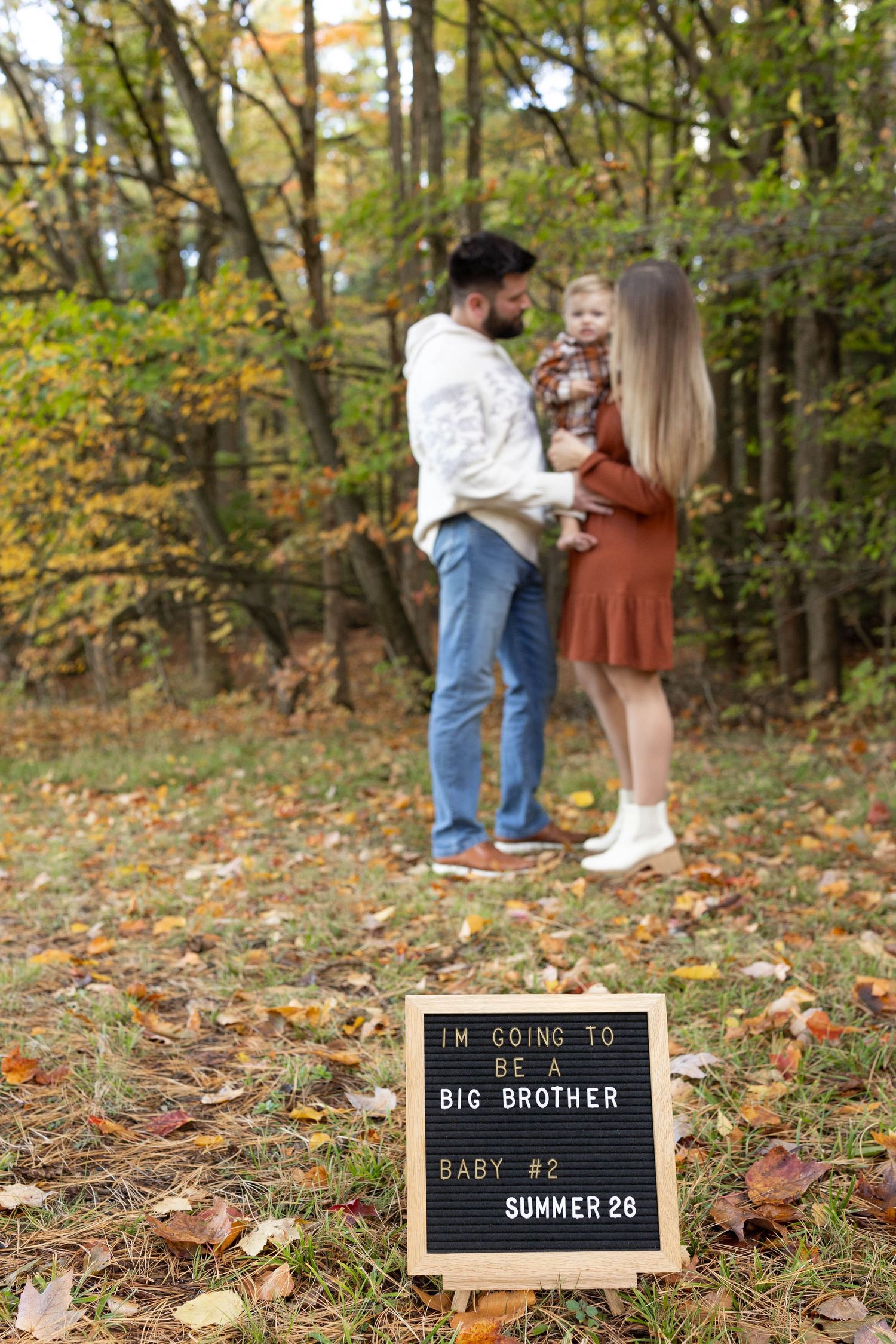A pregnancy announcement letterboard sits in autumn leaves while a family stands together in the wooded background.