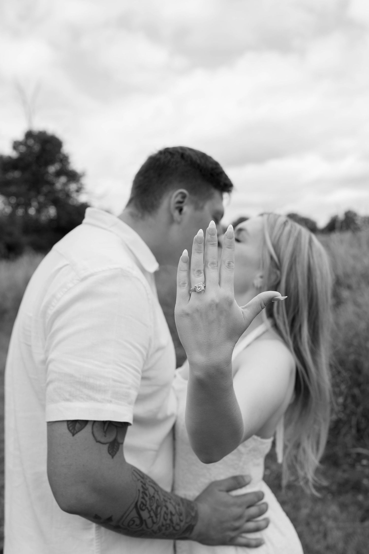 Black and white photo of a couple kissing outdoors, woman showing off engagement ring with tattooed man embracing her.