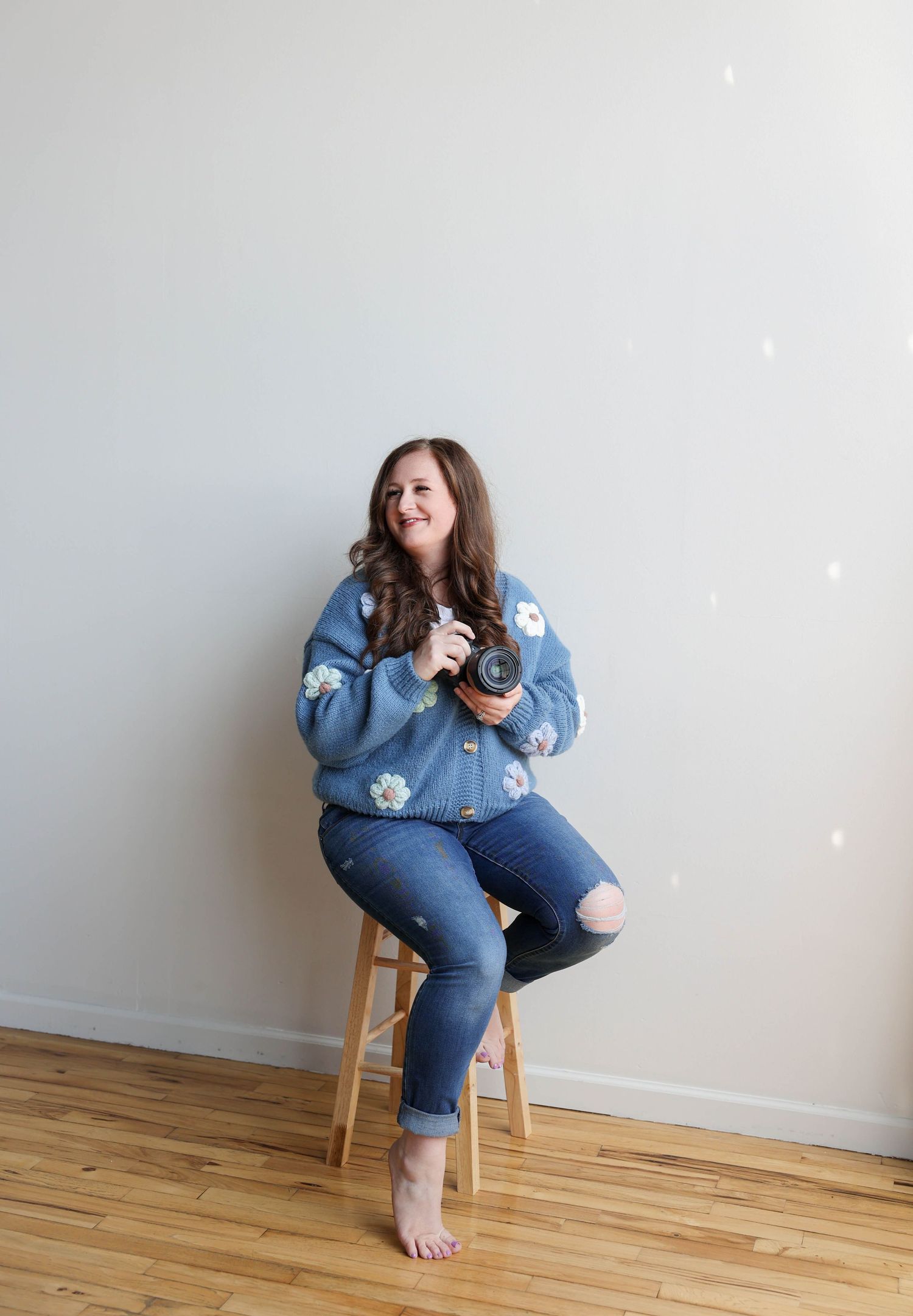 Woman in blue floral sweater and ripped jeans sits on stool holding camera, smiling in bright studio.