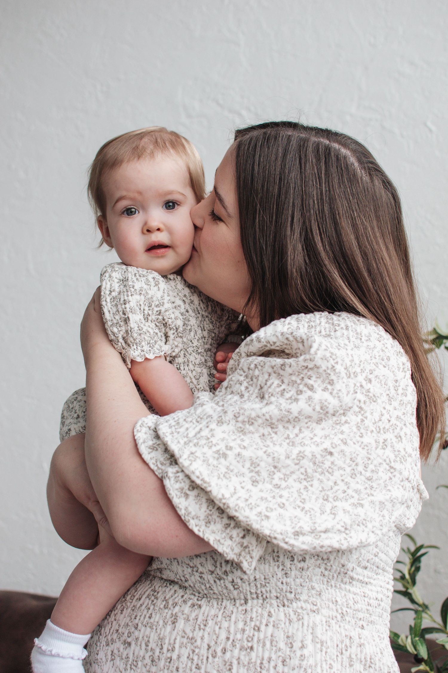 A tender moment shared in white matching outfits against a light gray wall with delicate greenery in the background.