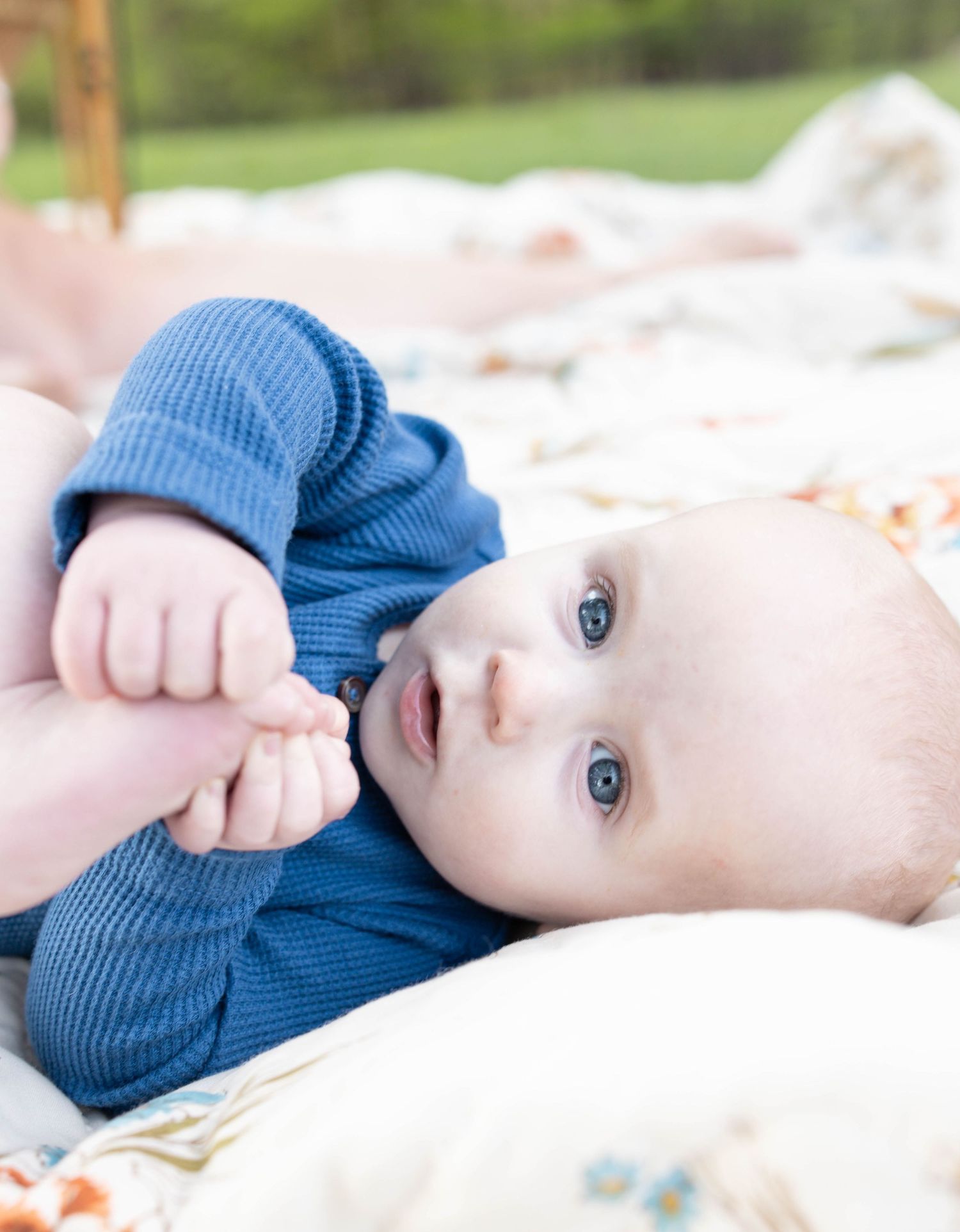 Baby wearing a blue knit sweater lying on a white blanket outdoors with bright eyes.