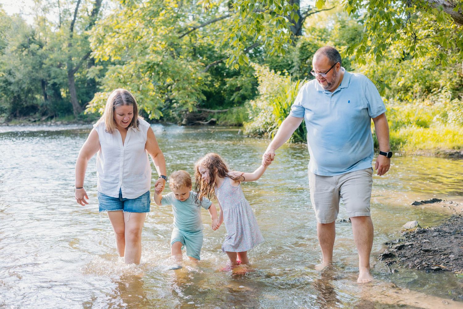 Family of four wading in a shallow river surrounded by lush green trees on a sunny day.