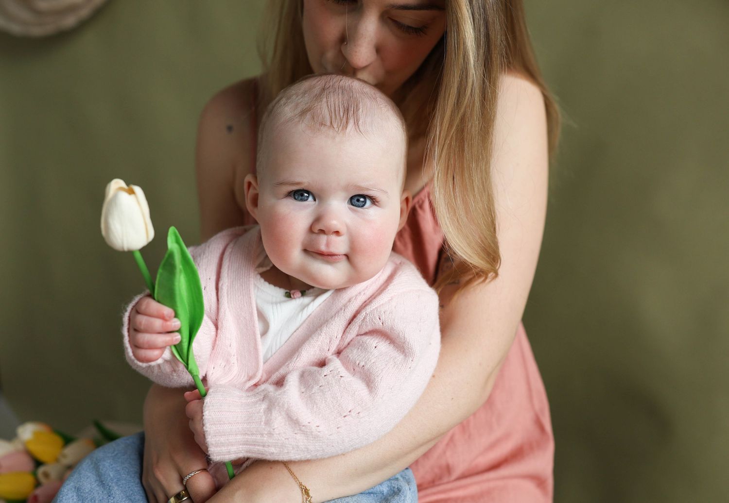 Mother kissing baby girl holding white tulip in pink cardigan.