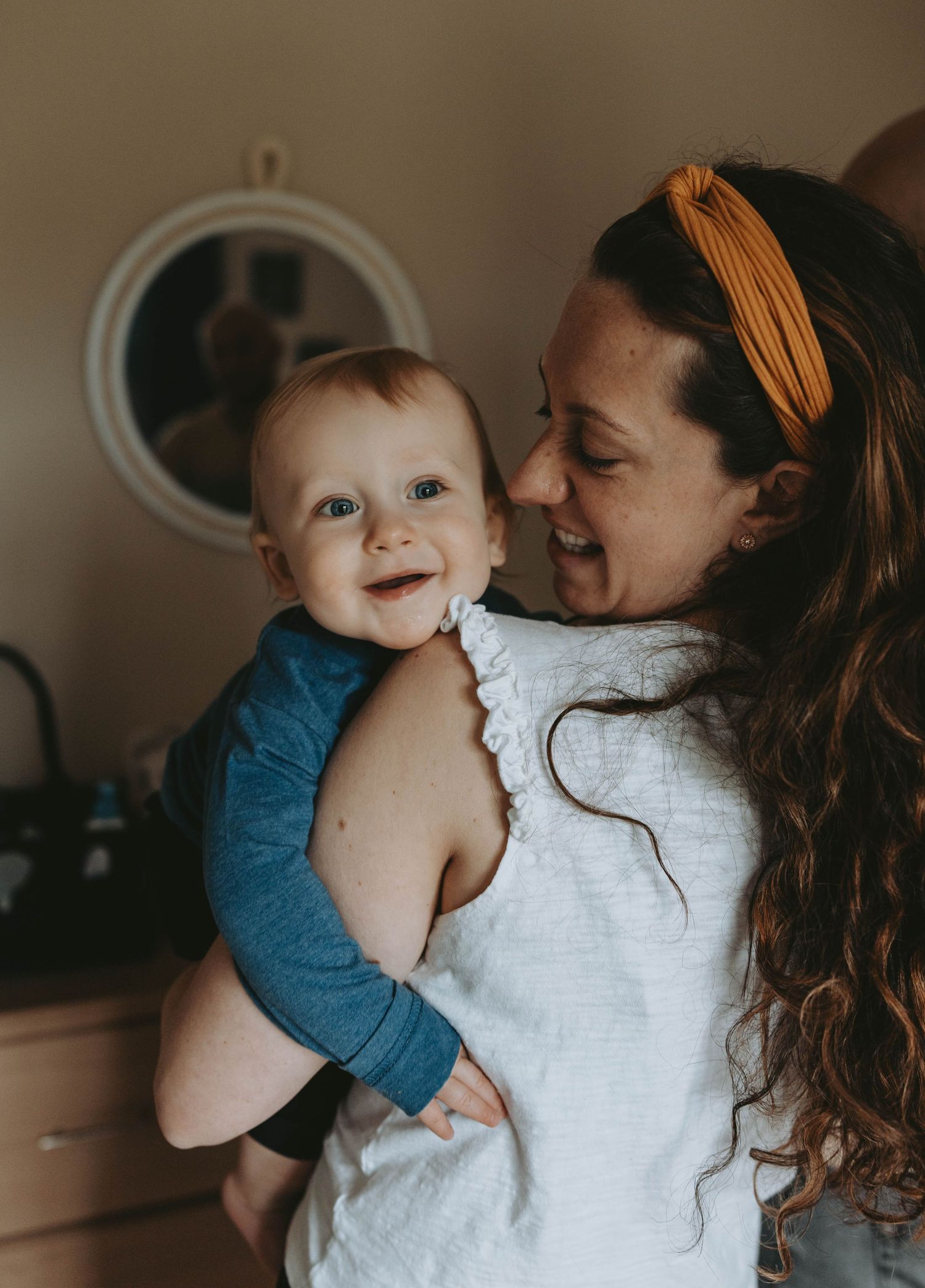 Smiling baby in blue outfit being held by a curly-haired mother wearing a yellow headband.