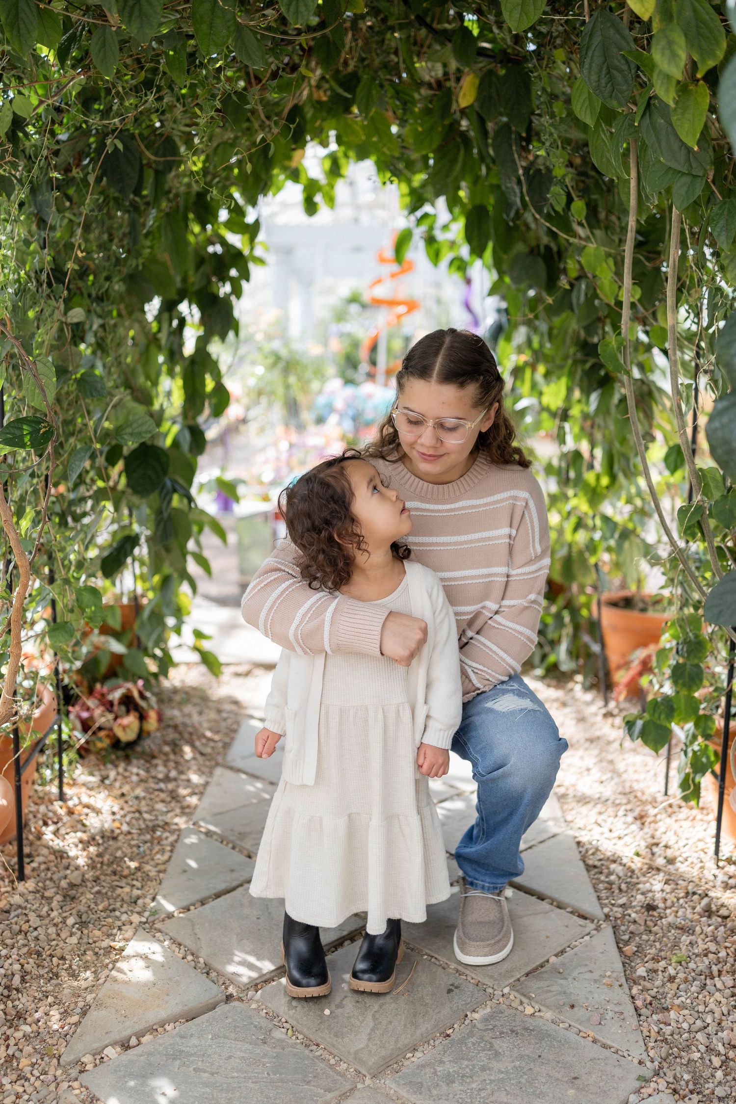 Parent and child share a sweet moment under a green garden archway dressed in matching neutral tones.