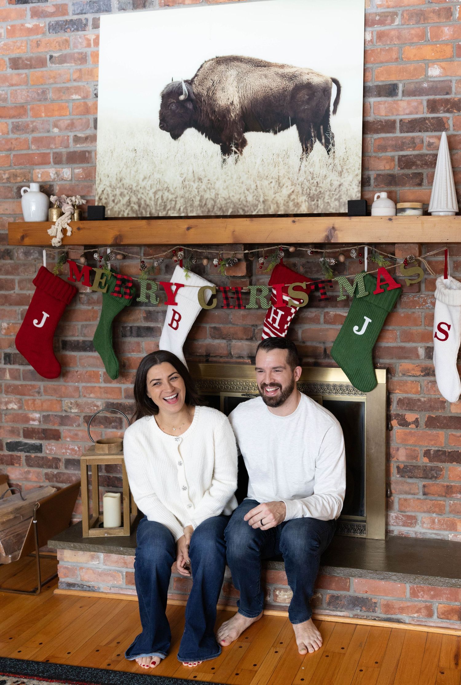 A couple sits on a wooden floor by a brick wall decorated with Christmas stockings and festive holiday decor.